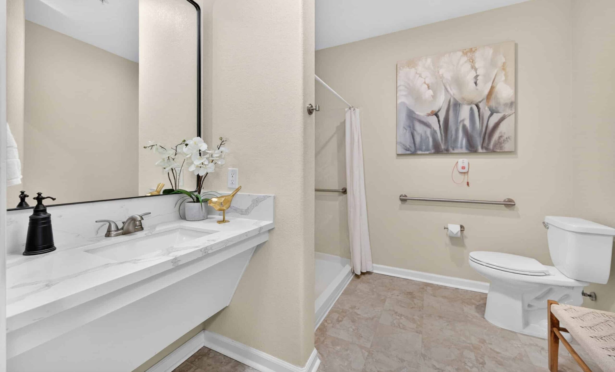 Accessible bathroom in Eskaton Granite Bay senior living features a marble sink, mirror, toilet, walk-in shower with grab bars, beige walls, tiled floor, and floral artwork. A wooden chair sits by the wall for added comfort.