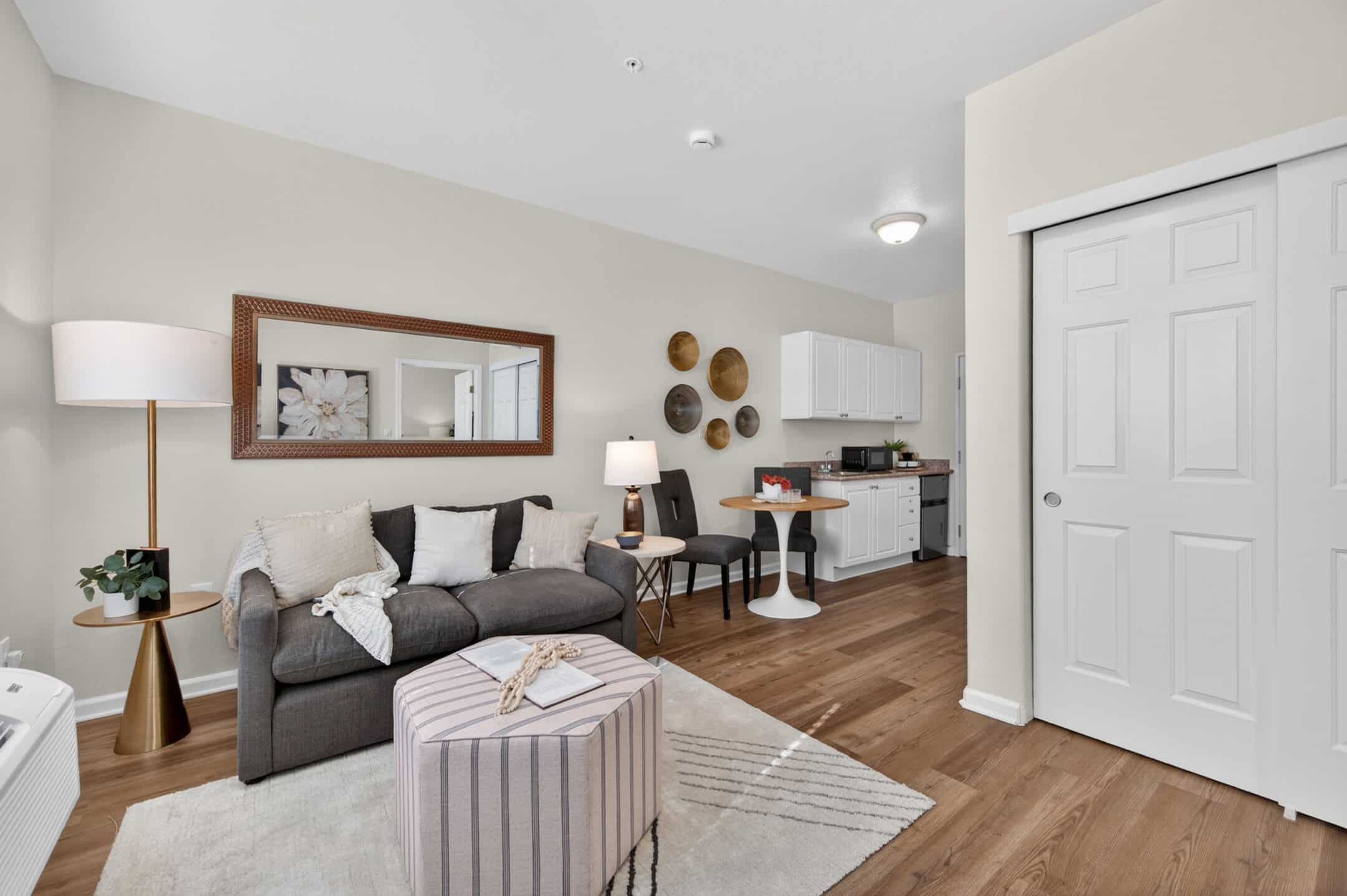 A modern, cozy living space with a gray sofa, pillows, and throw blanket offers versatile living options; features include a round ottoman, floor lamp, small dining table for two, wall decor, wood flooring, and a compact kitchen in the background.