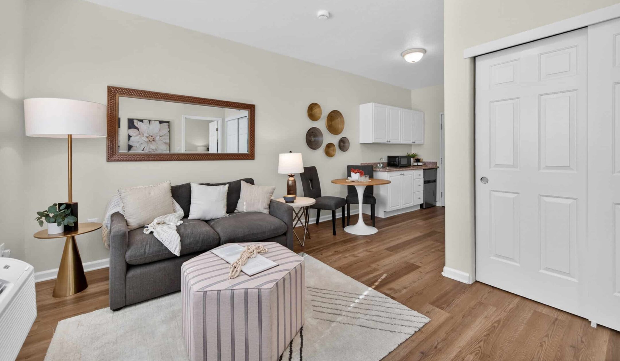 A modern, cozy living space with a gray sofa, pillows, and throw blanket offers versatile living options; features include a round ottoman, floor lamp, small dining table for two, wall decor, wood flooring, and a compact kitchen in the background.