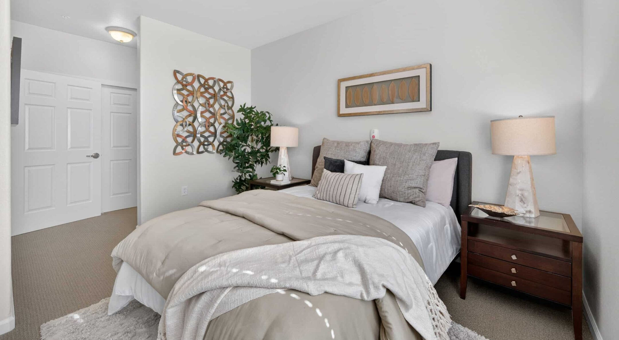A modern bedroom with a neatly made bed, neutral bedding, two bedside tables with lamps, wall art, and a green plant highlights stylish living options. The room is bright and well-lit, with a closed white door in the background.