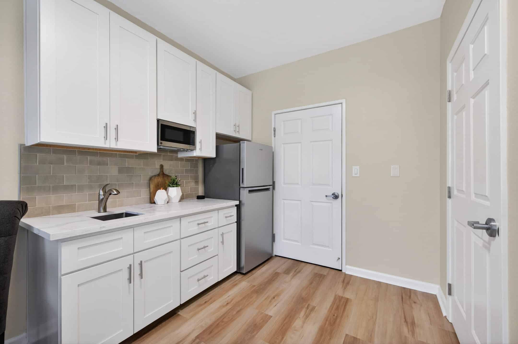 A modern kitchenette in Eskaton Granite Bay senior living features white cabinets, a stainless steel refrigerator, microwave, sink, and light wood flooring. Green subway tile backsplash and minimal decor create a clean, neutral look.