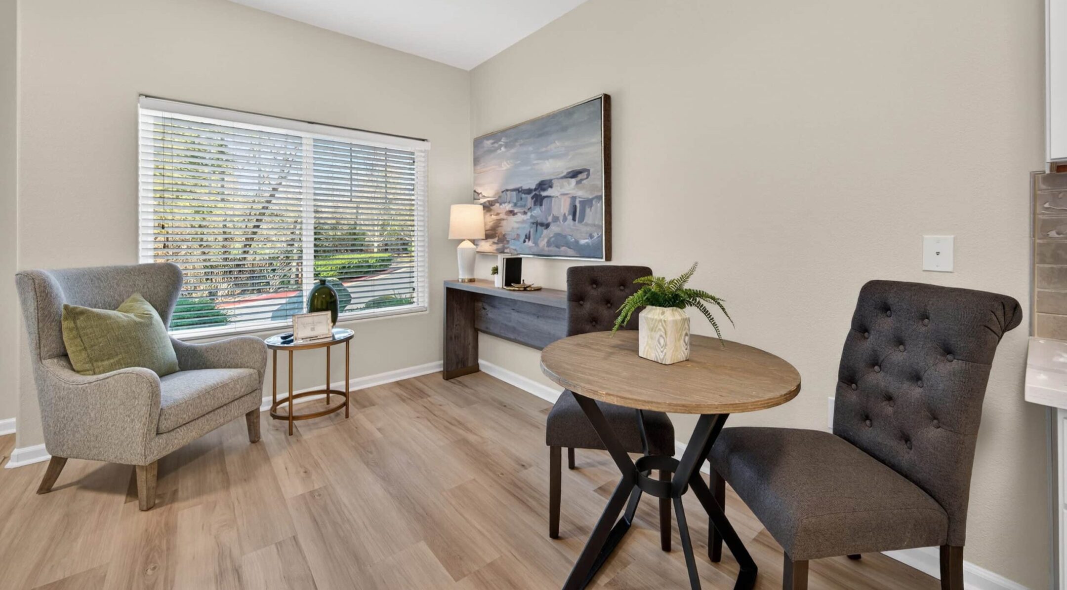 A bright, modern room with wood floors offers versatile living options—featuring a round table with two dark chairs, a cozy armchair by the window, a side table with decor, and a wall-mounted desk with a lamp and artwork.