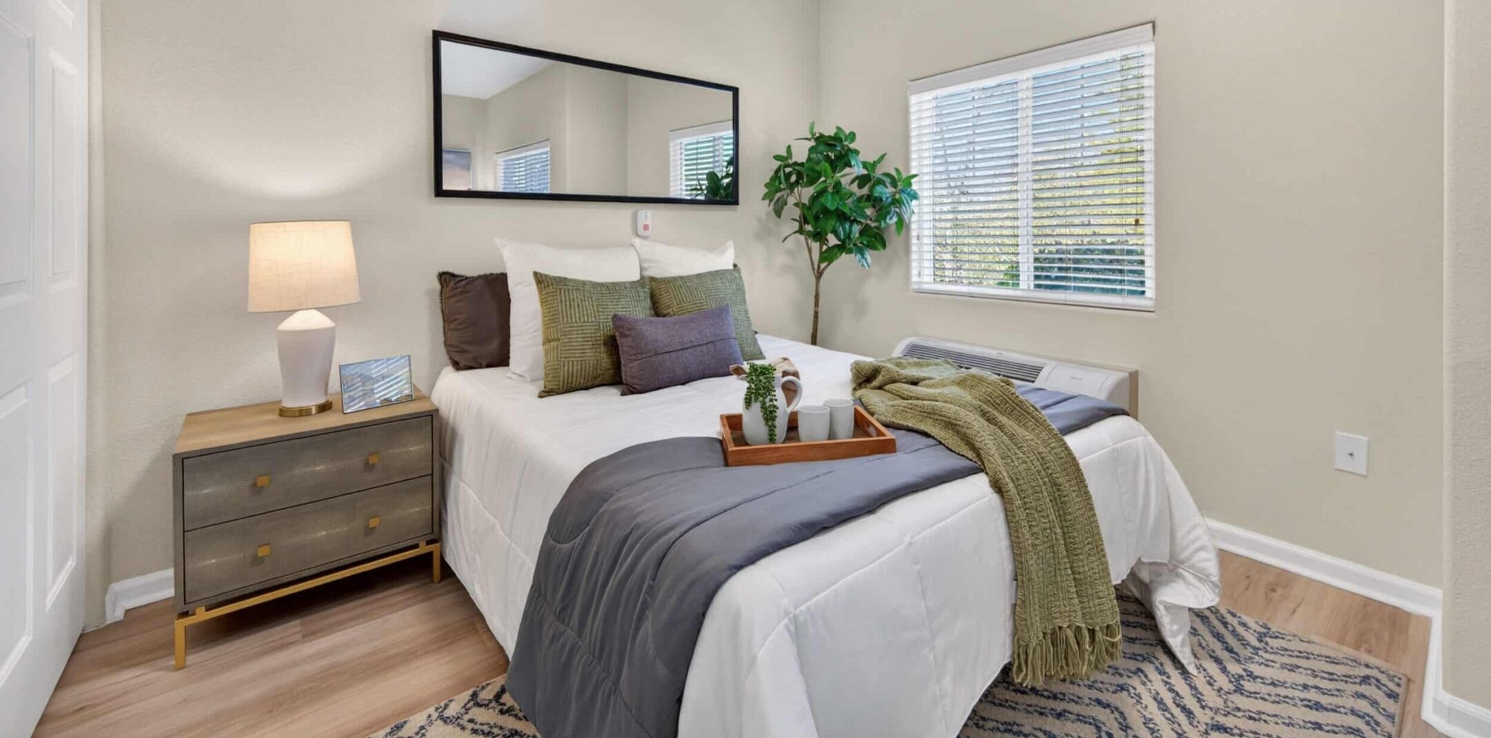 A cozy bedroom with a neatly made bed, green and gray pillows, a green throw, a bedside table with a lamp, a potted plant, a large mirror, and a window with blinds letting in natural light.