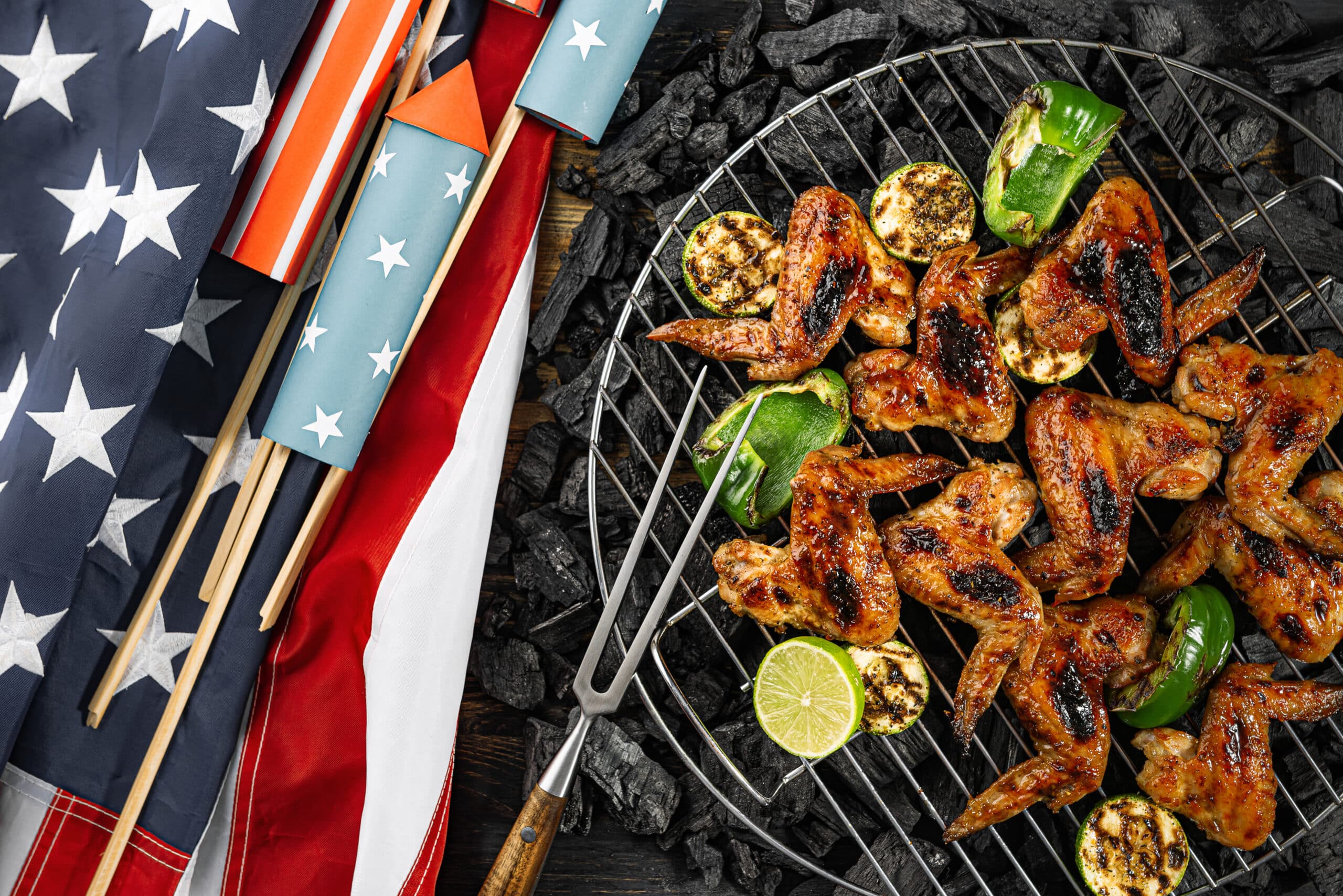 Grilled chicken wings, zucchini, and lime sizzle on a charcoal grill at a Stars & Stripes BBQ, set beside an American flag and festive red, white, and blue fireworks rockets.