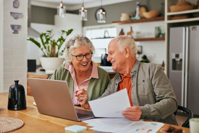 Smiling elderly couple sits at a kitchen table in their Eskaton Granite Bay independent living apartment, looking at a laptop and holding papers together, appearing happy and engaged.