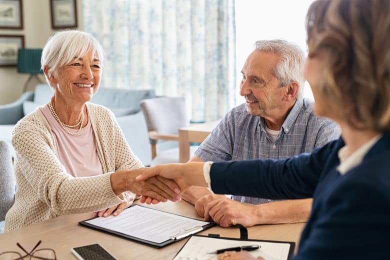 An older woman smiles and shakes hands with another person across a table, while an older man sits beside her, also smiling. A clipboard with papers and pen is on the table, suggesting a meeting or agreement—perhaps at Eskaton Granite Bay assisted living, where residents are welcomed with warmth and support.