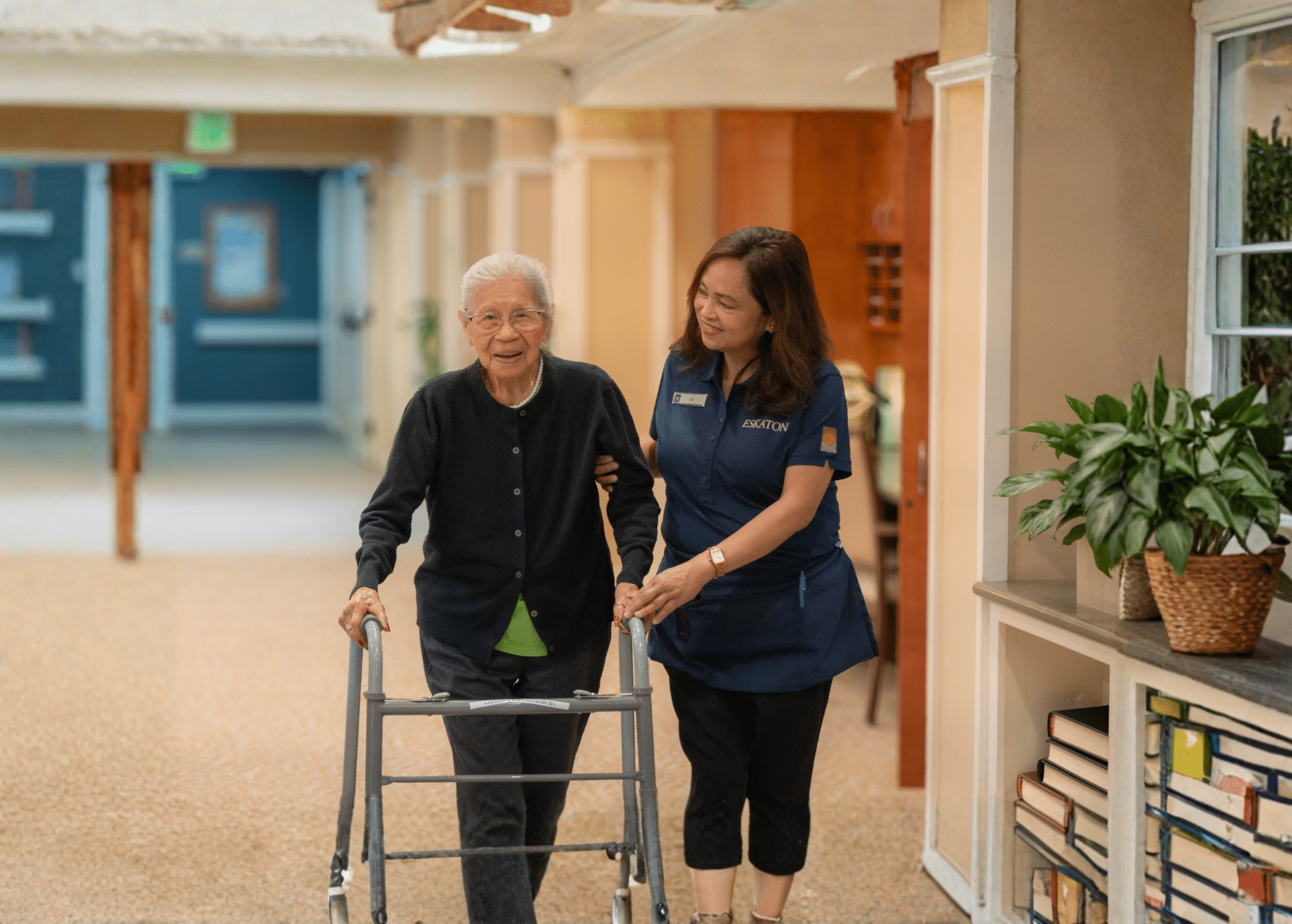 An elderly woman using a walker is assisted by a caregiver in a hallway with warm lighting. Books and a plant are on a shelf nearby.