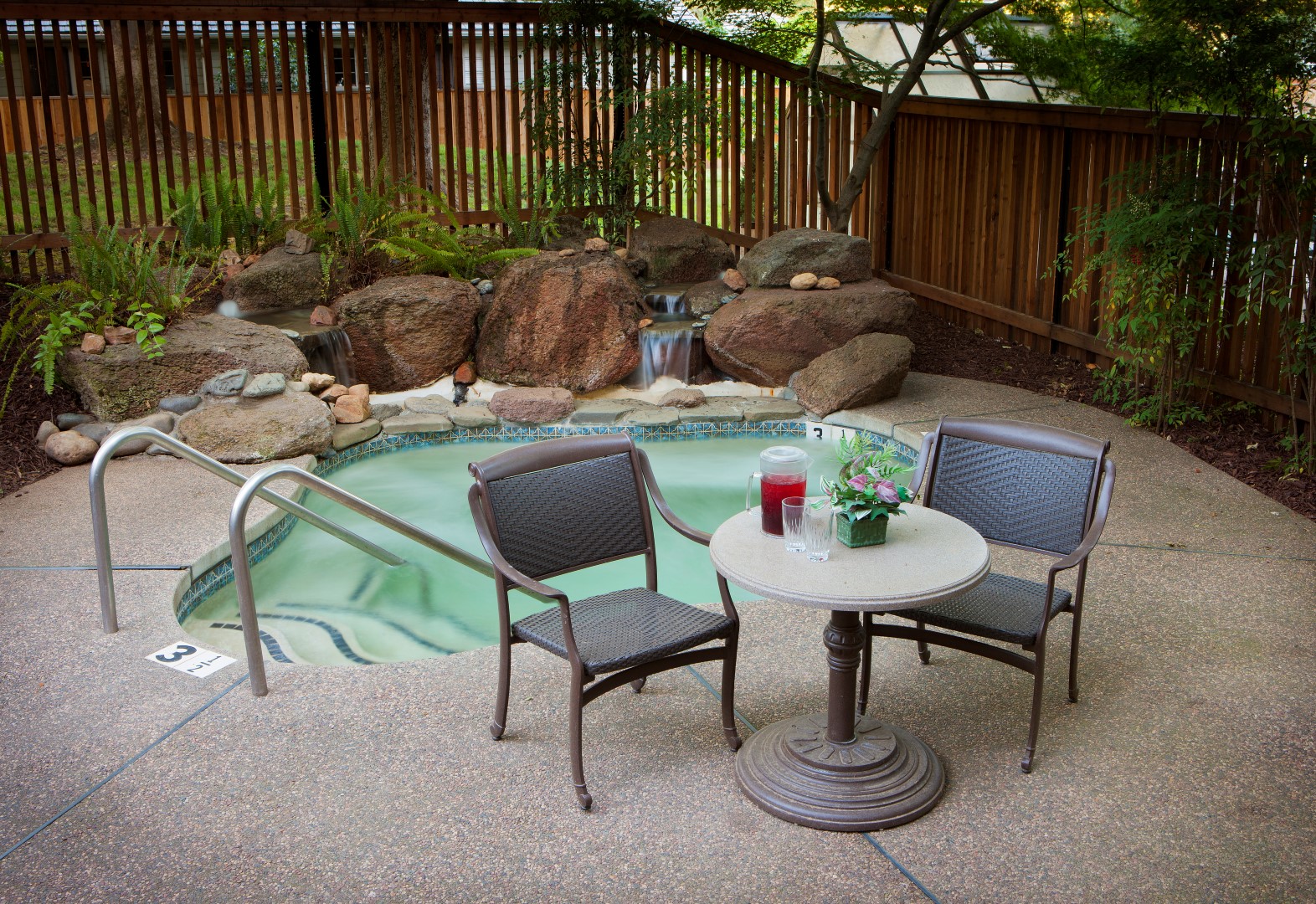 A small backyard hot tub with a waterfall feature is surrounded by rocks and plants. Two chairs and a round table with drinks and a potted plant sit on the patio next to the water. A wooden fence encloses the area.