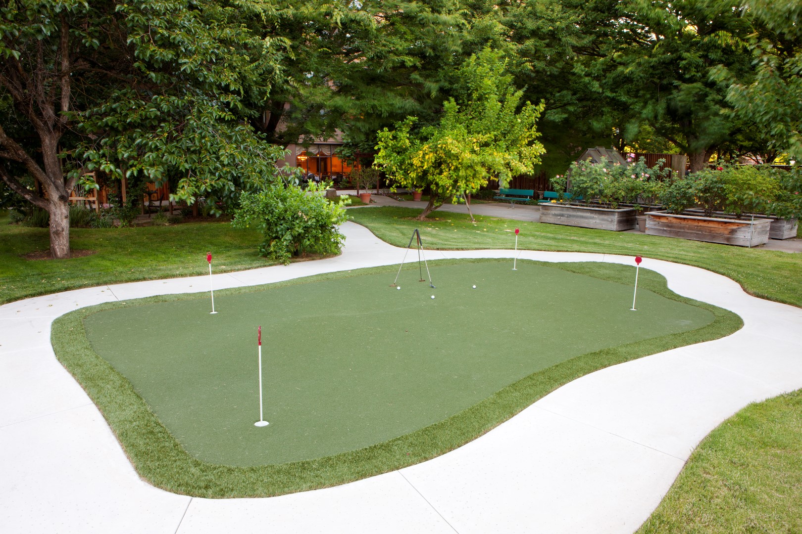 A small outdoor putting green with four golf holes, surrounded by white concrete paths and lush green trees and plants on a sunny day. Raised garden beds are visible in the background.