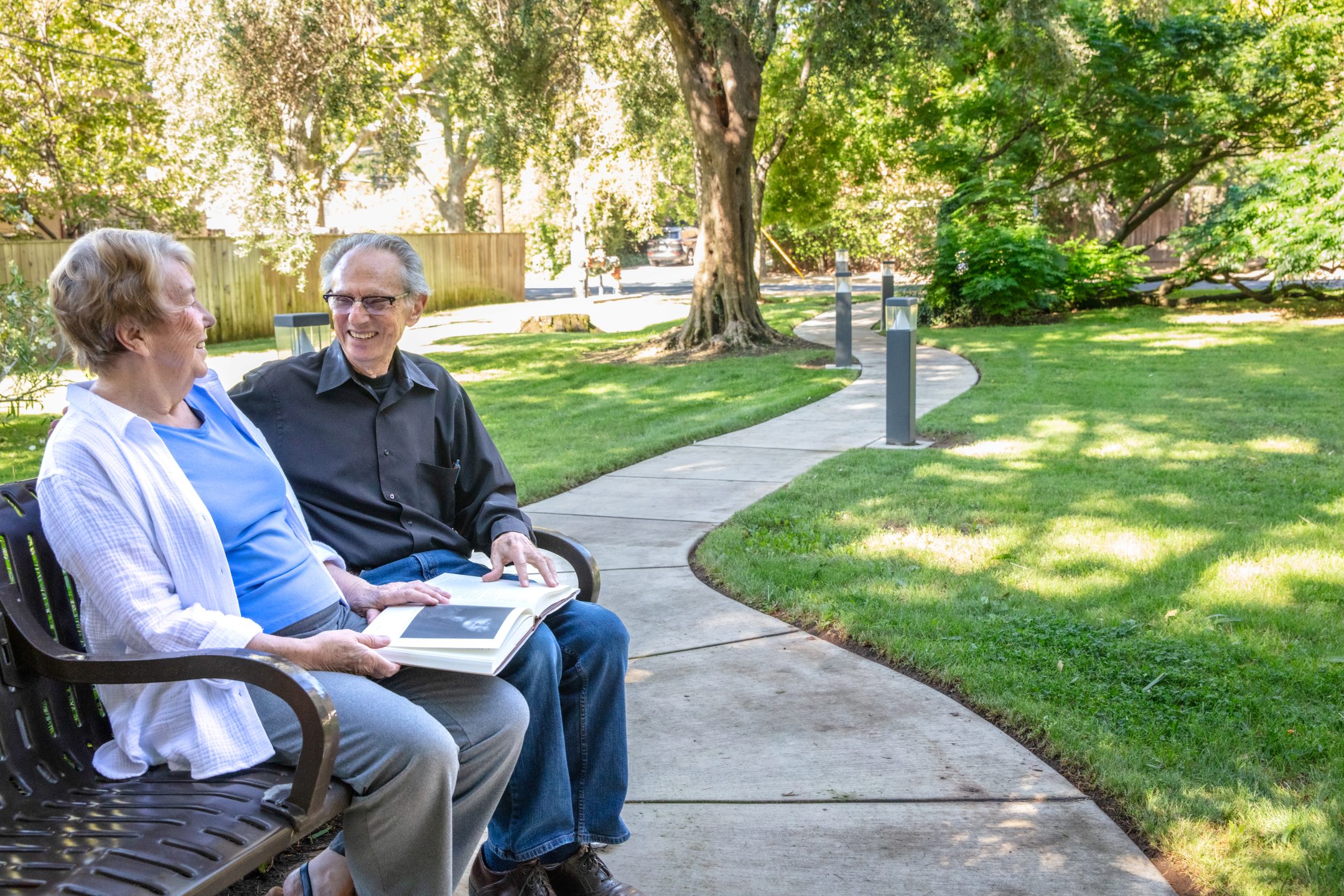 An older woman and man sit on a bench in Eskaton Land Park, smiling and talking. The woman holds an open photo album on her lap, while a winding path and green trees create a peaceful background.