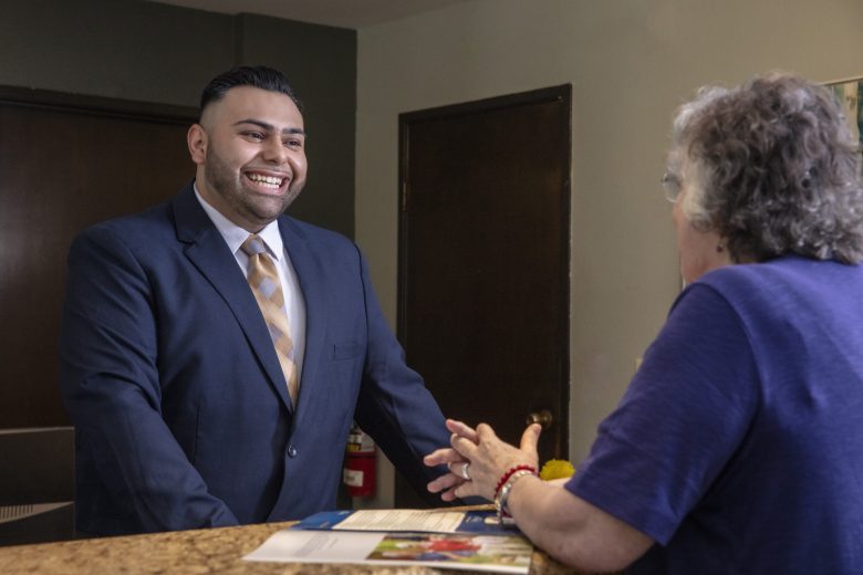A smiling man in a suit stands behind a counter, engaging in conversation with an older woman in a blue top as they discuss documents and brochures—offering a glimpse into the welcoming lifestyle at Eskaton Land Park.