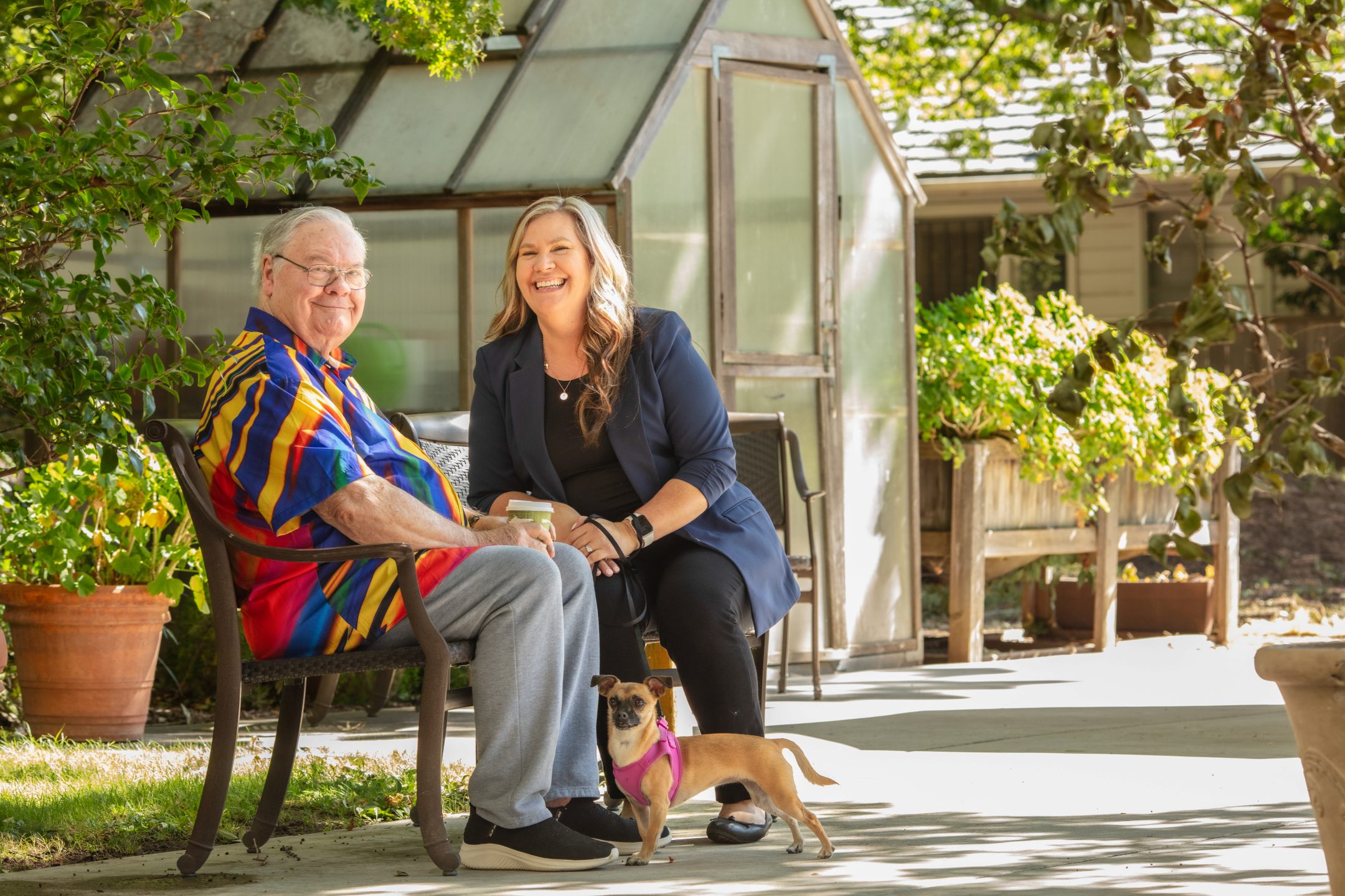 An elderly man in a colorful shirt sits on a bench next to a smiling woman with a coffee cup. A small dog stands at their feet. They are outdoors near a greenhouse, enjoying the lush greenery and nearby amenities.
