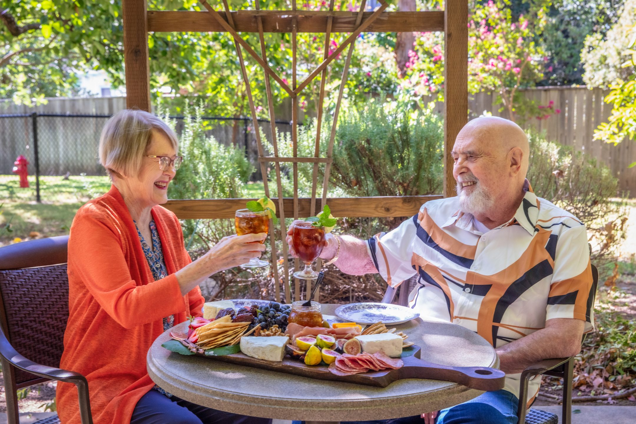 An older woman and man sit at a patio table in a garden, smiling and clinking glasses of iced tea. Their Land Park Dining experience features a charcuterie board with cheese, crackers, fruit, and meats amid lush greenery.