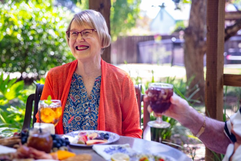 An older woman with short hair and glasses, wearing an orange cardigan and patterned blue top, smiles while sitting at an outdoor table in an Independent Living community, enjoying a sunny day with food, drinks, and good company.