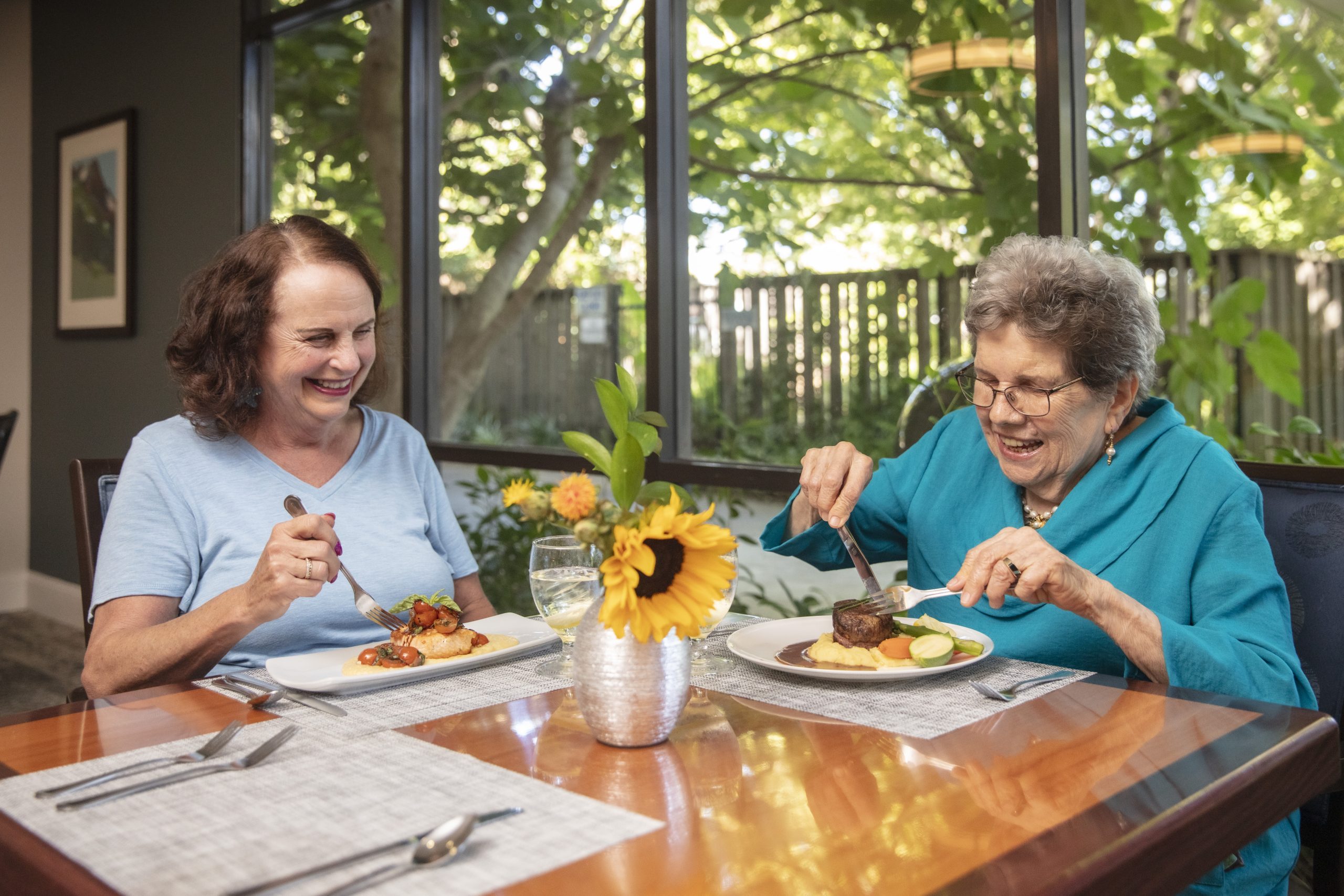 Two older women sit at a dining table by a window at Eskaton Land Park, enjoying a meal together. Both are smiling and engaged in pleasant conversation. A vase with a sunflower decorates the table, with trees and greenery outside—showcasing the lifestyle at Eskaton Land Park.