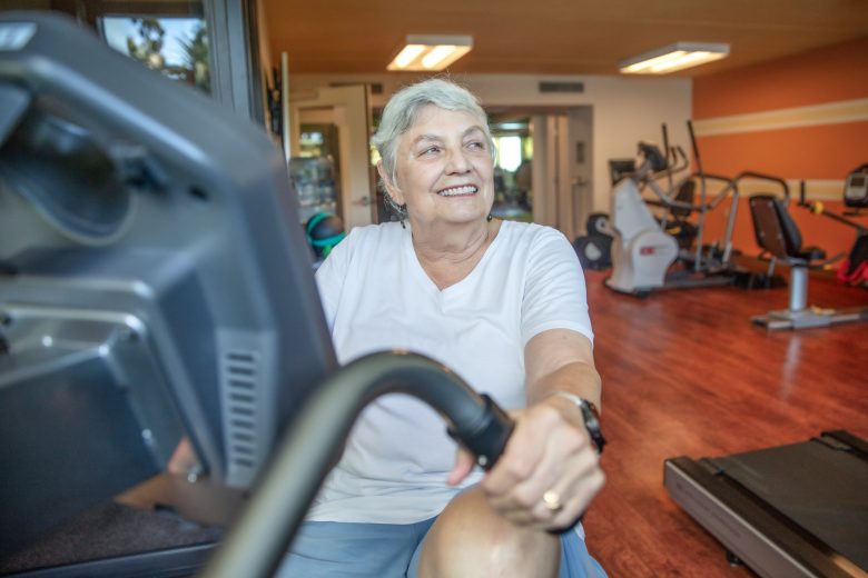 An older woman in a white shirt smiles while using an exercise bike, enjoying the resident experience in a gym filled with various fitness equipment and wooden floors.