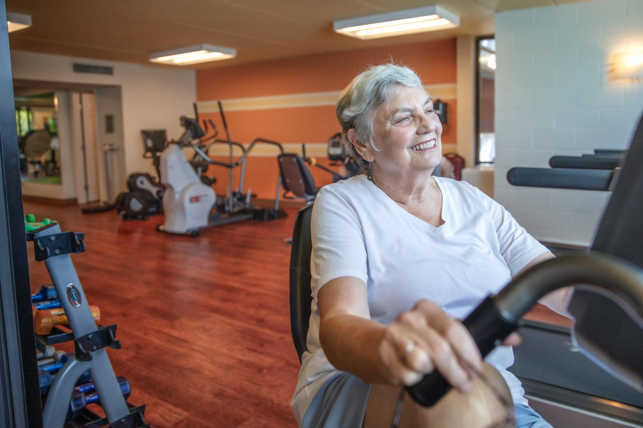 An older woman with gray hair and a white shirt smiles while using an exercise bike in a brightly lit fitness room featuring modern amenities, various workout equipment, and wooden floors.