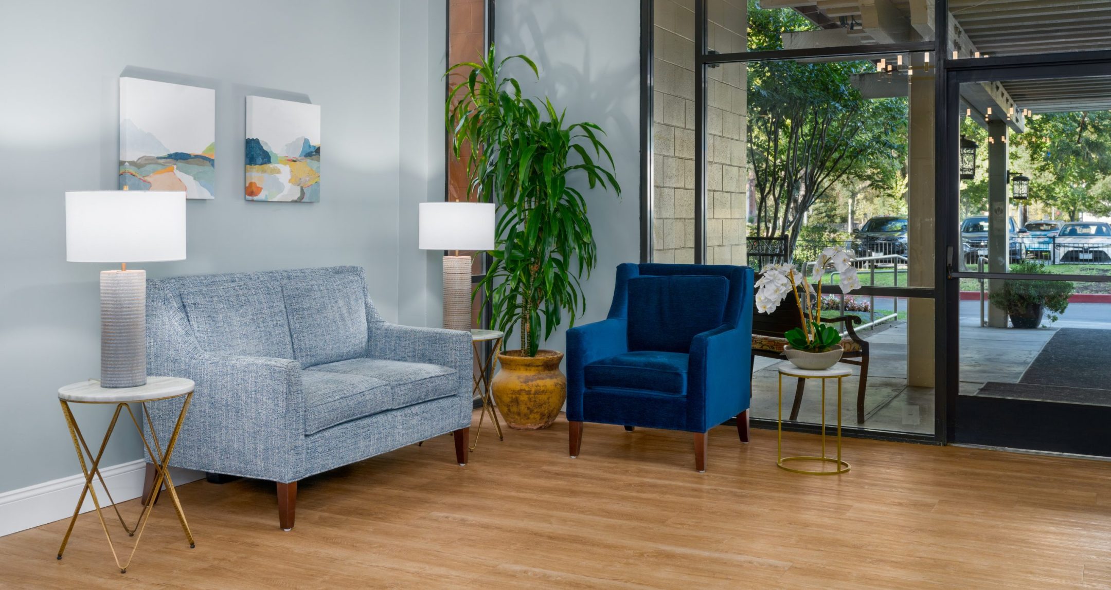 A modern Independent Living waiting area features a blue loveseat, navy armchair, two side tables with lamps, a potted plant, wall art, and large windows revealing trees and parked cars outside.