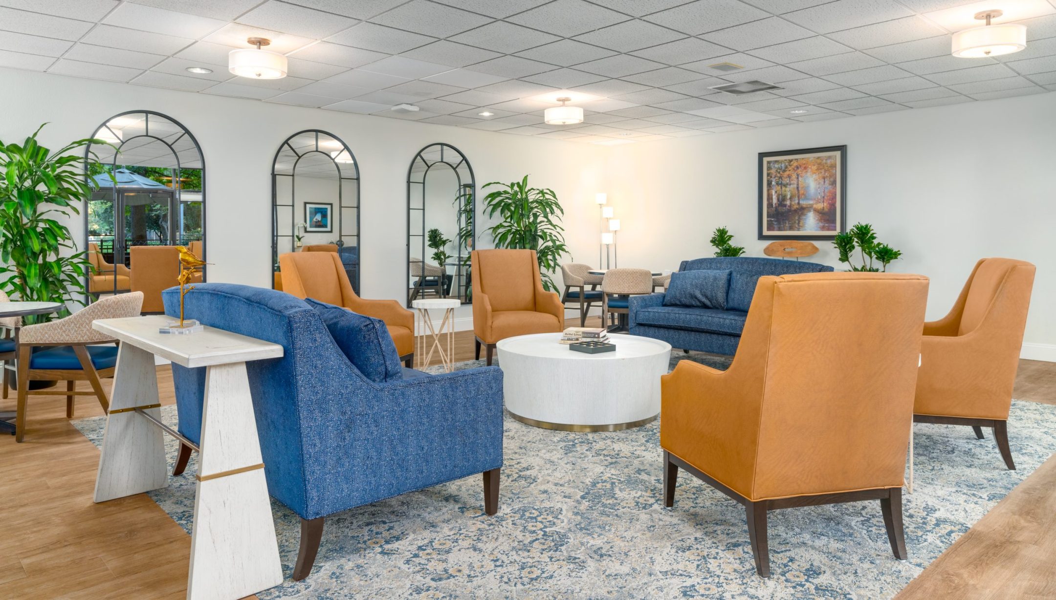 A modern lounge at Eskaton Land Park features blue sofas, orange armchairs, a round white coffee table, large mirrors, green plants, wall art, and soft lighting on a patterned blue rug with wood flooring.