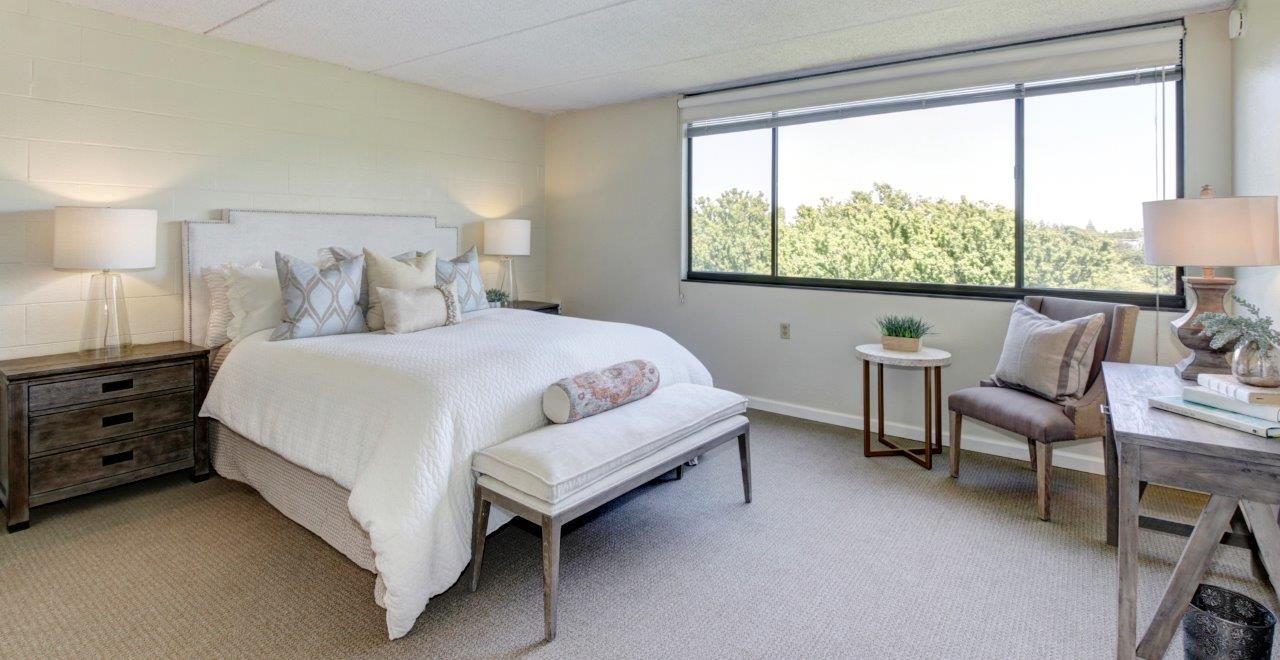 Bright, modern bedroom with a large window, a neatly made bed with pillows, two bedside tables with lamps, a bench at the foot of the bed, and a cozy seating area with a chair, side table, and decorative items.