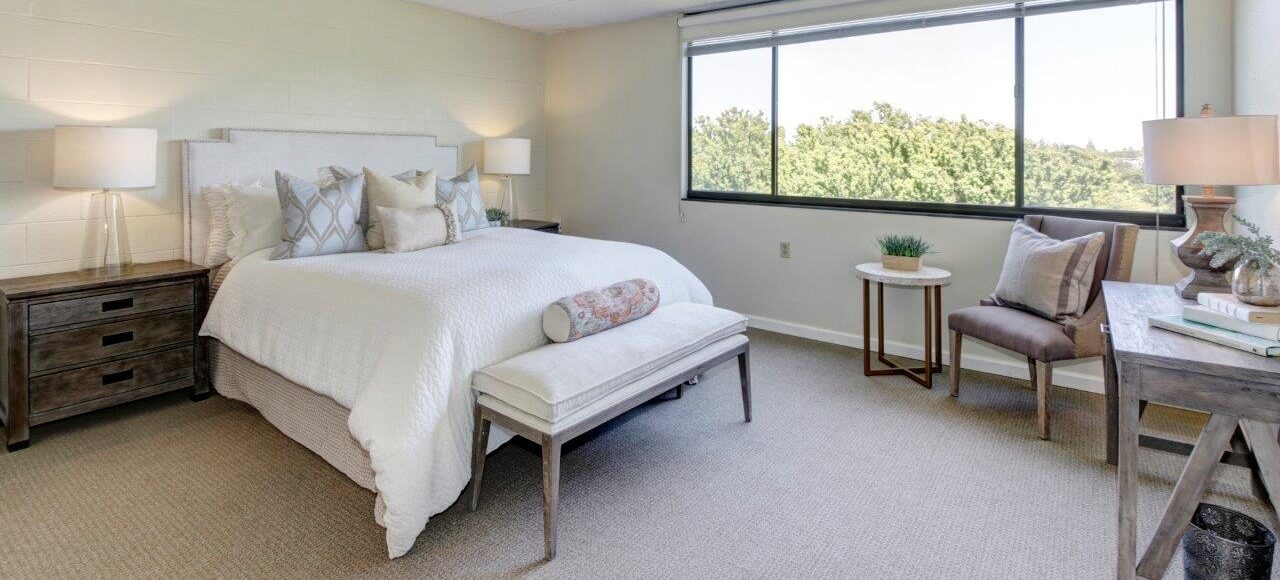 Bright, modern bedroom with a large window, a neatly made bed with pillows, two bedside tables with lamps, a bench at the foot of the bed, and a cozy seating area with a chair, side table, and decorative items.