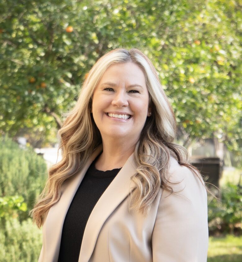 A woman with long, wavy blonde hair smiles while standing outdoors in front of green trees at Eskaton Land Park independent living. She is wearing a tan blazer over a black top and is illuminated by natural sunlight.