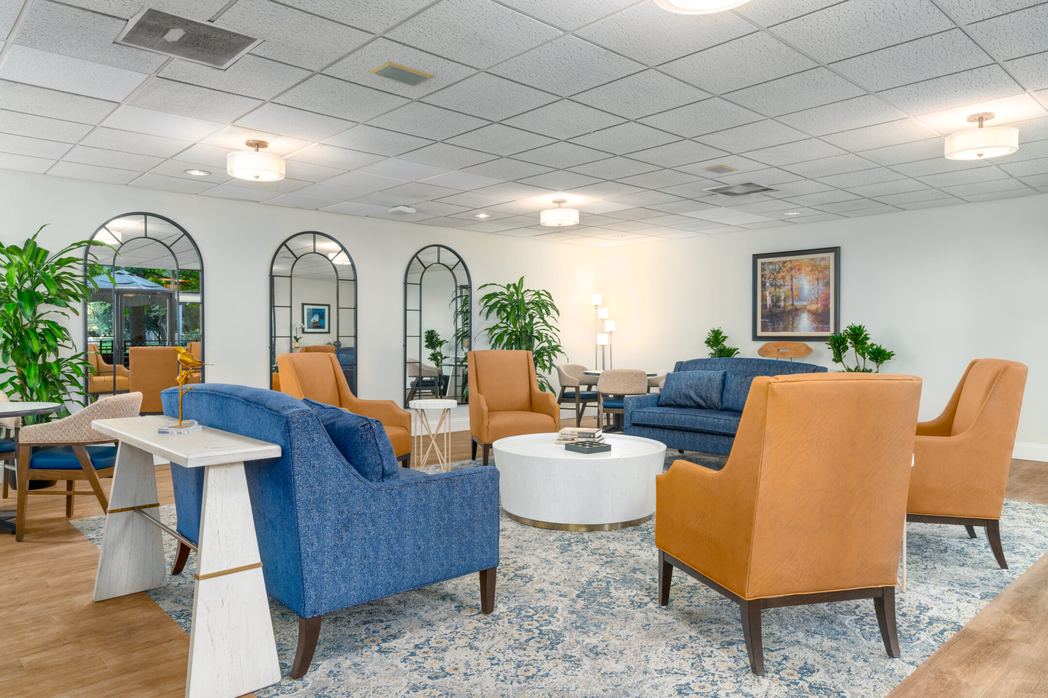 A modern lounge with blue and tan armchairs, a blue sofa, white round coffee table, large mirrors, indoor plants, and soft ceiling lights creating a bright atmosphere—download a brochure to explore this inviting space.