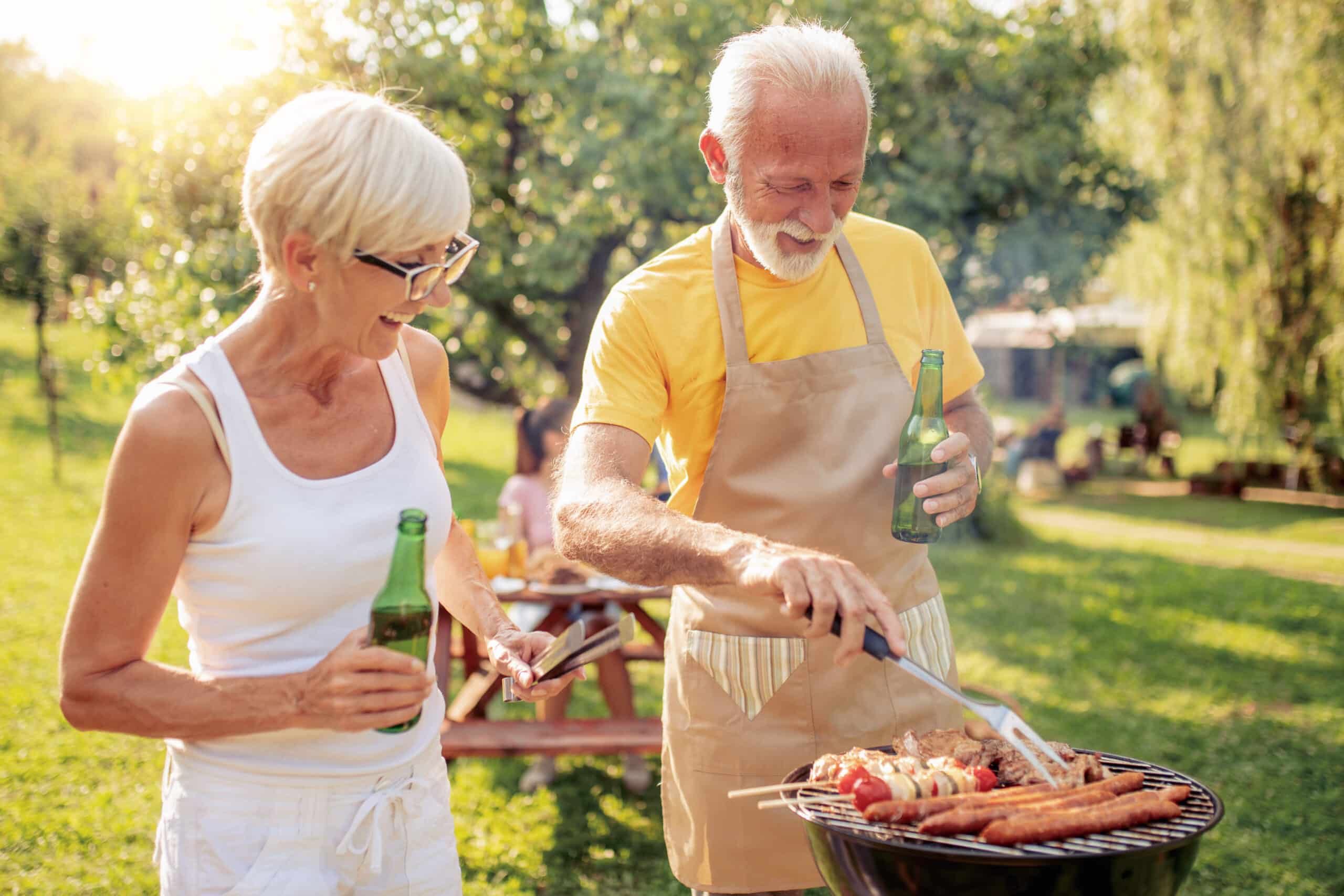 An older couple enjoys a sunny outdoor barbecue, smiling and holding drinks as the man grills sausages and vegetables on a round grill in a green backyard.