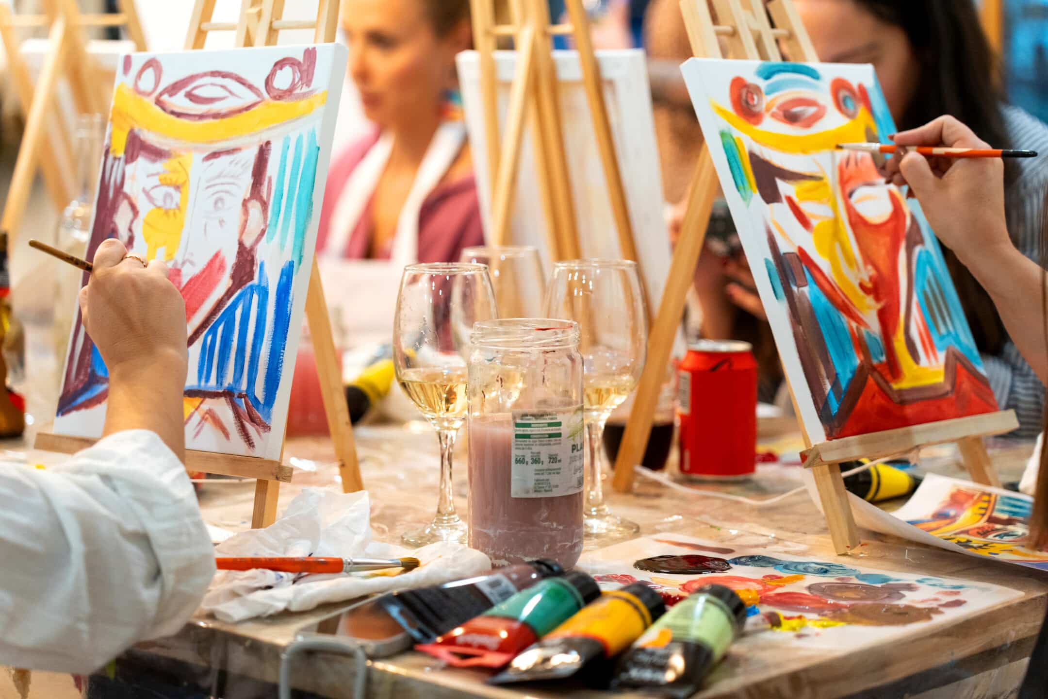 People painting colorful abstract portraits at a table with easels, paint tubes, brushes, and wine glasses. The scene is lively, with unfinished paintings and art supplies scattered around.