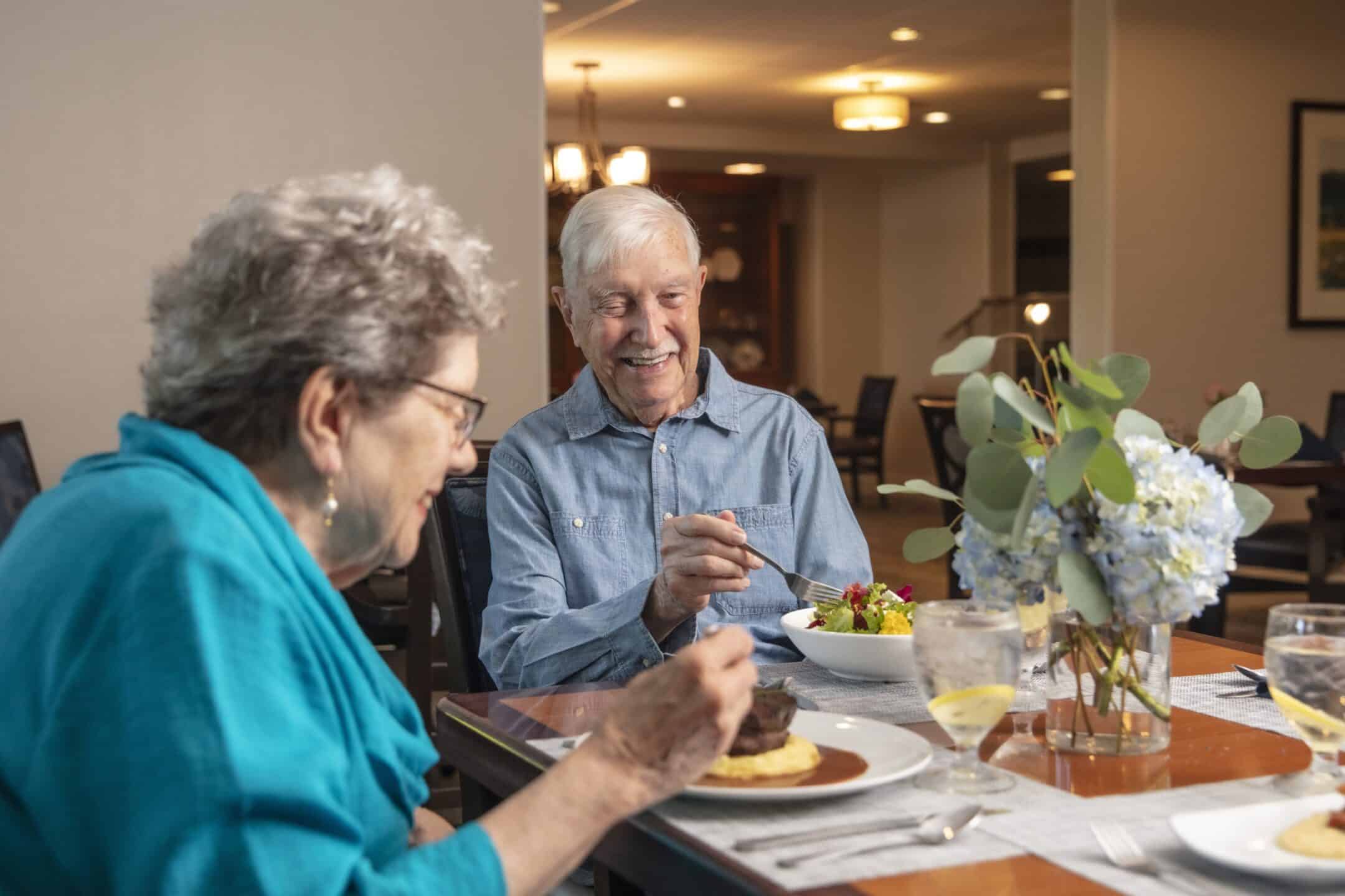 Two older adults sit at a dining table in a restaurant, smiling and enjoying a meal as part of a Learning Over Lunch event. A vase with blue flowers and glasses of water are on the table. The background is softly lit, creating a warm atmosphere—perfect for conversations about topics like Downsizing Made Simple.