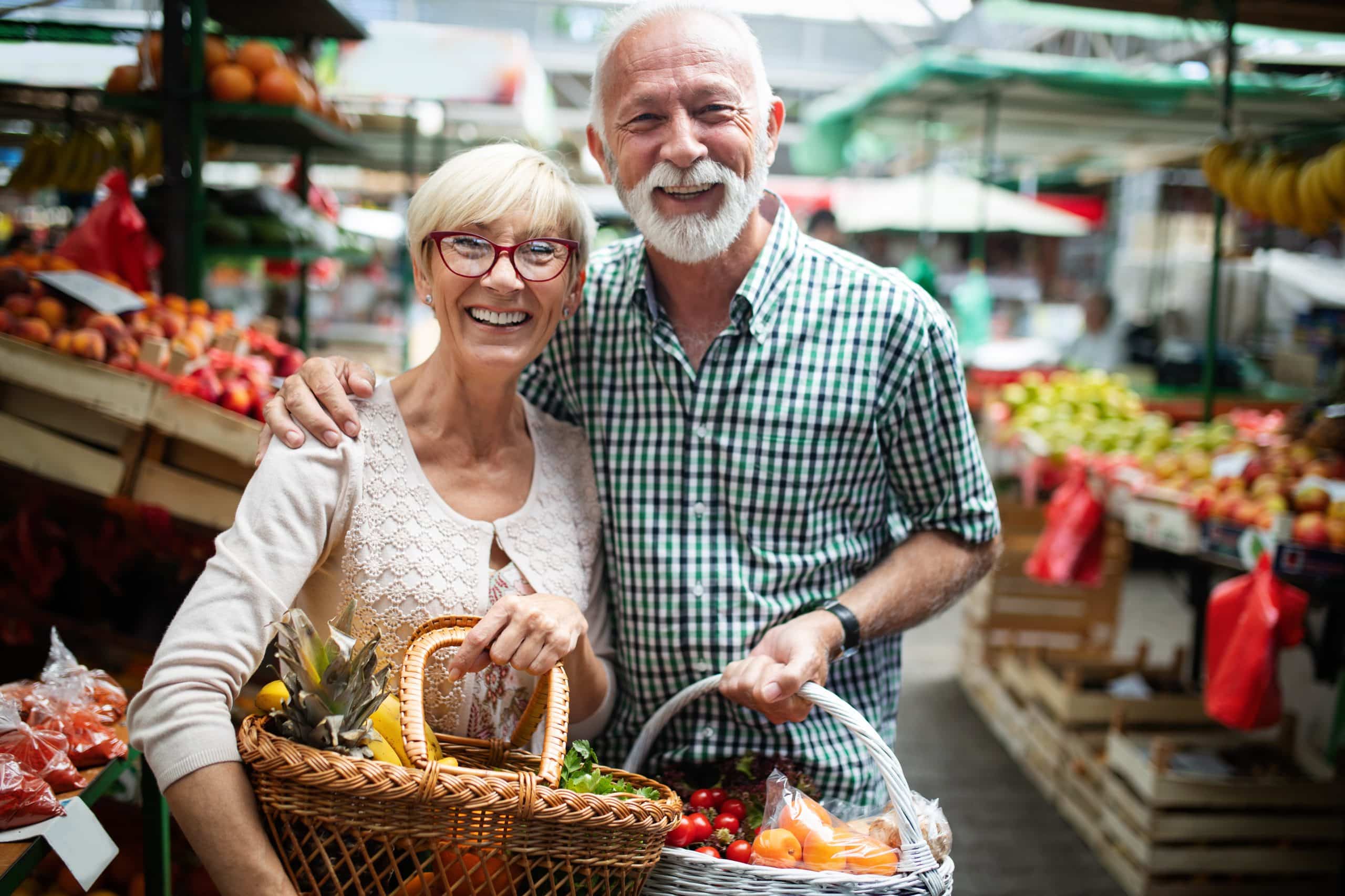 Smiling older couple holding baskets of fresh produce, standing together at an outdoor Farmer’s Market surrounded by colorful fruits and vegetables.