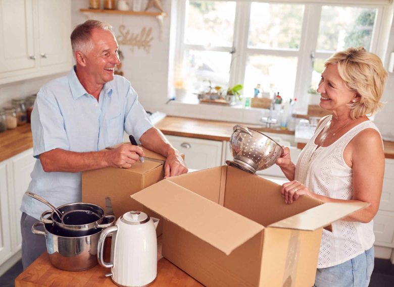 Smiling older couple in a kitchen packing kitchenware into cardboard boxes, preparing to move or organize for Independent Living. The man writes on a box while the woman holds a metal colander. Sunlight streams through the window behind them.