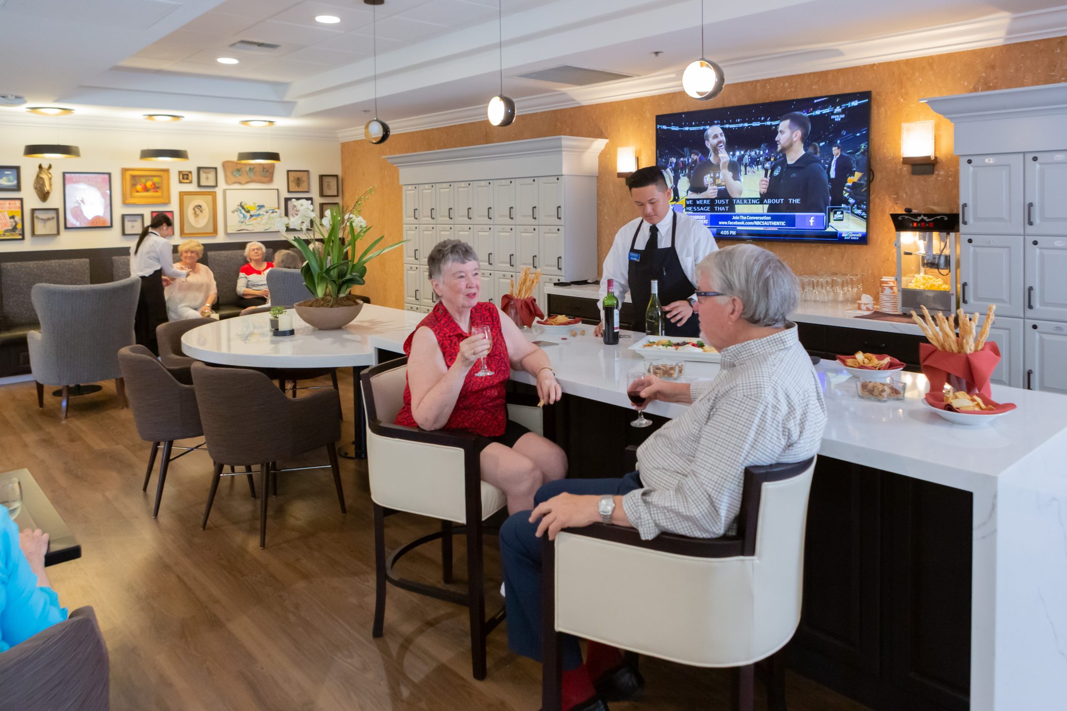 Seniors enjoy drinks and conversation at a modern lounge bar with snacks on the counter, while a bartender serves wine. Other guests relax in the background, and a TV plays sports news.