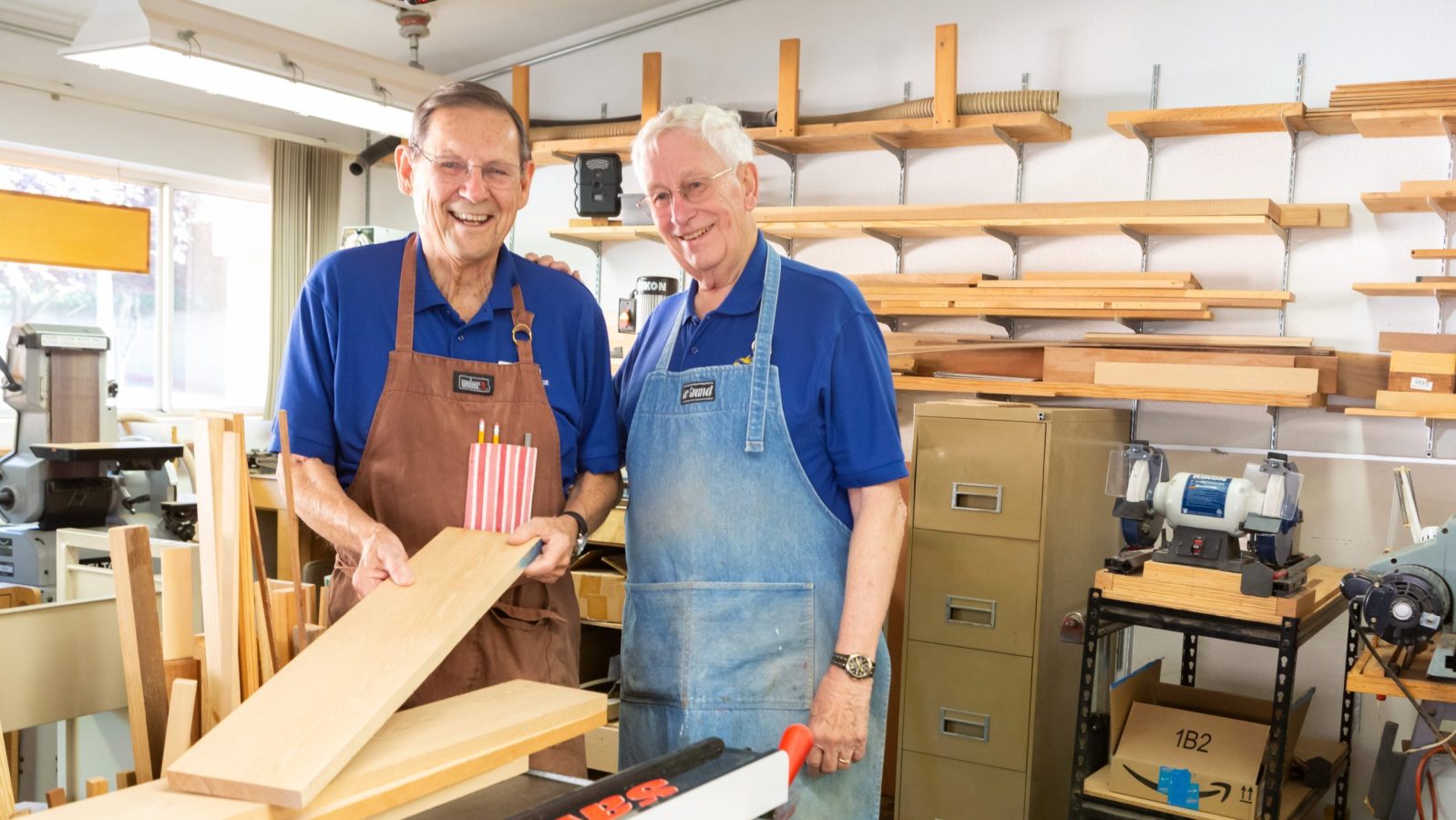 Two older men stand smiling in a woodworking shop within a Life Plan Community. One holds wooden boards and pencils, while the other stands beside him. Both wear aprons, surrounded by shelves, planks, and woodworking tools.