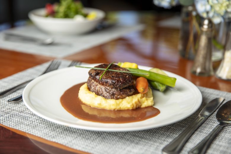 A plated dish featuring a seared filet mignon on mashed potatoes with brown sauce, garnished with carrots, green beans, and a chive, set on a dining table with utensils and a blurred salad in the background.