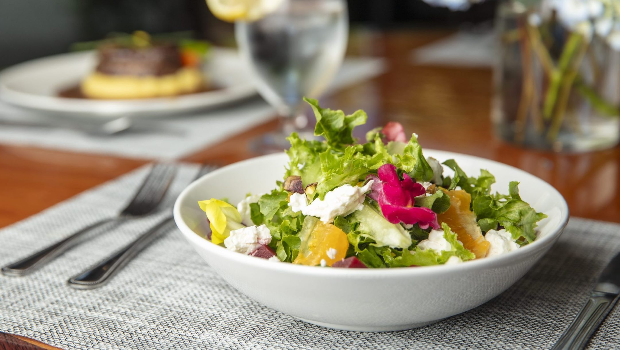 A white bowl filled with a fresh salad of leafy greens, orange slices, cheese, and edible flowers offers a vibrant culinary experience. It sits on a placemat with cutlery, alongside a glass of water and another meal blurred in the background.
