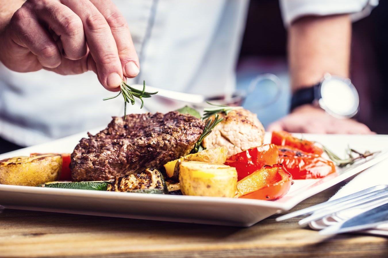 A chef showcases their culinary experience by garnishing a plated meal with fresh rosemary. The dish features grilled steak, roasted potatoes, zucchini, and bell peppers on a white square plate beside silverware on a wooden table.