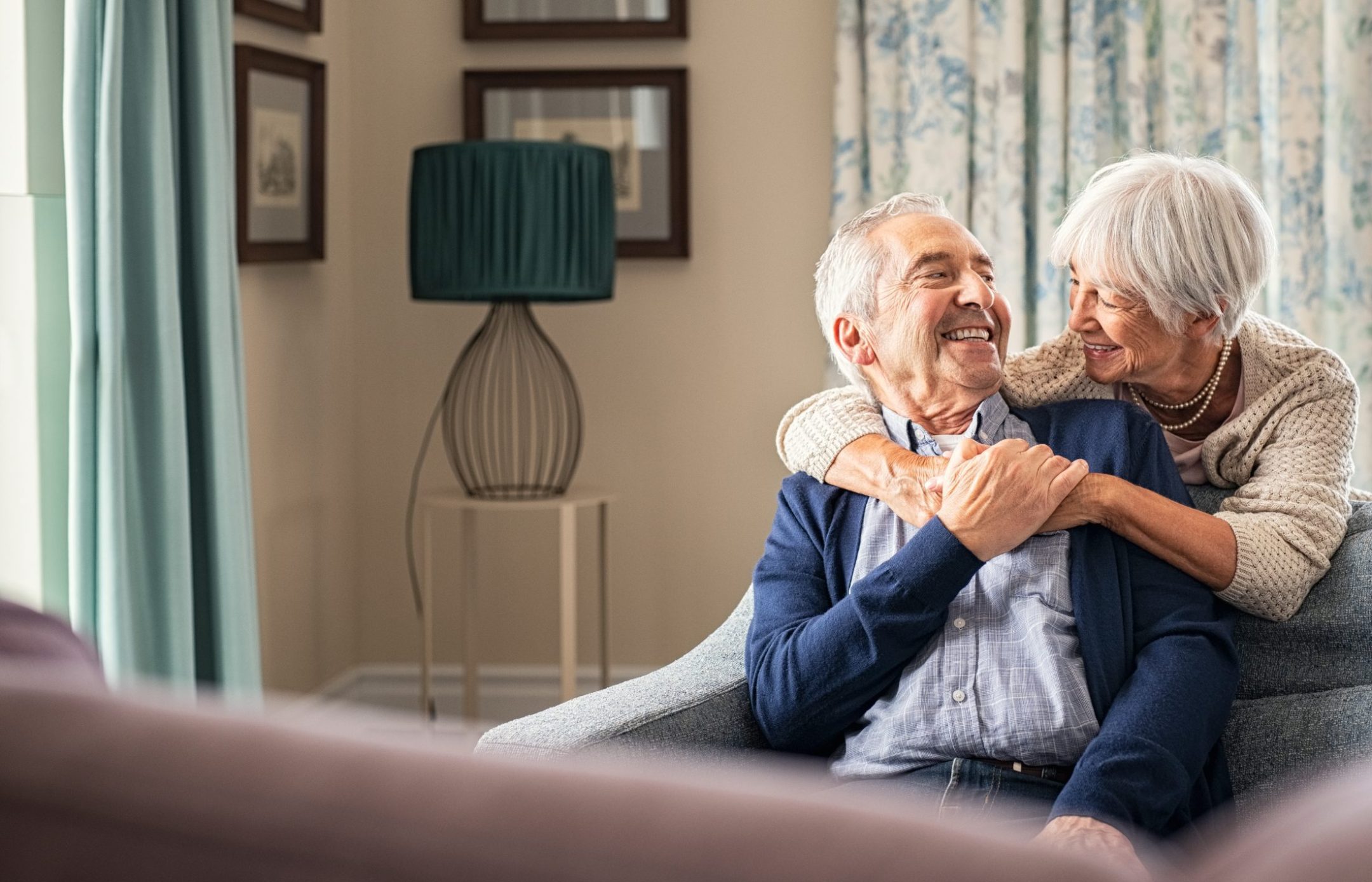 An elderly couple sits on a couch at home, smiling and embracing warmly, with the woman hugging the man from behind. The room is softly lit with patterned curtains and framed pictures on the wall.