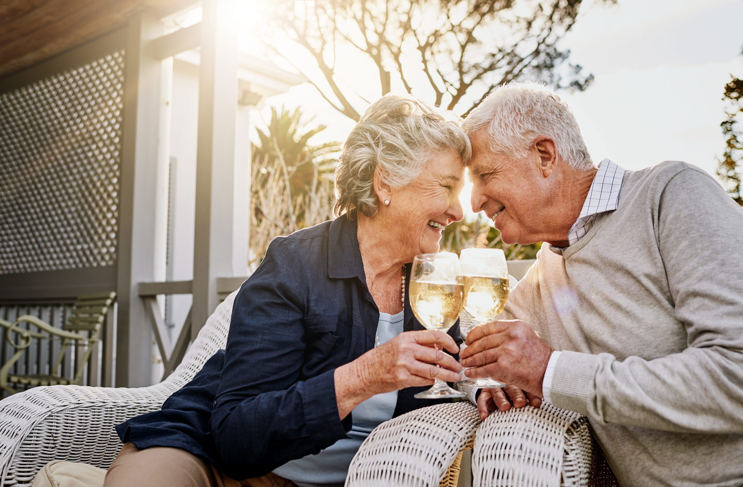 An elderly couple sits close together on outdoor chairs, smiling and touching foreheads while holding glasses of sparkling wine, enjoying a sunny day on the porch—celebrating the joys of Independent Living.