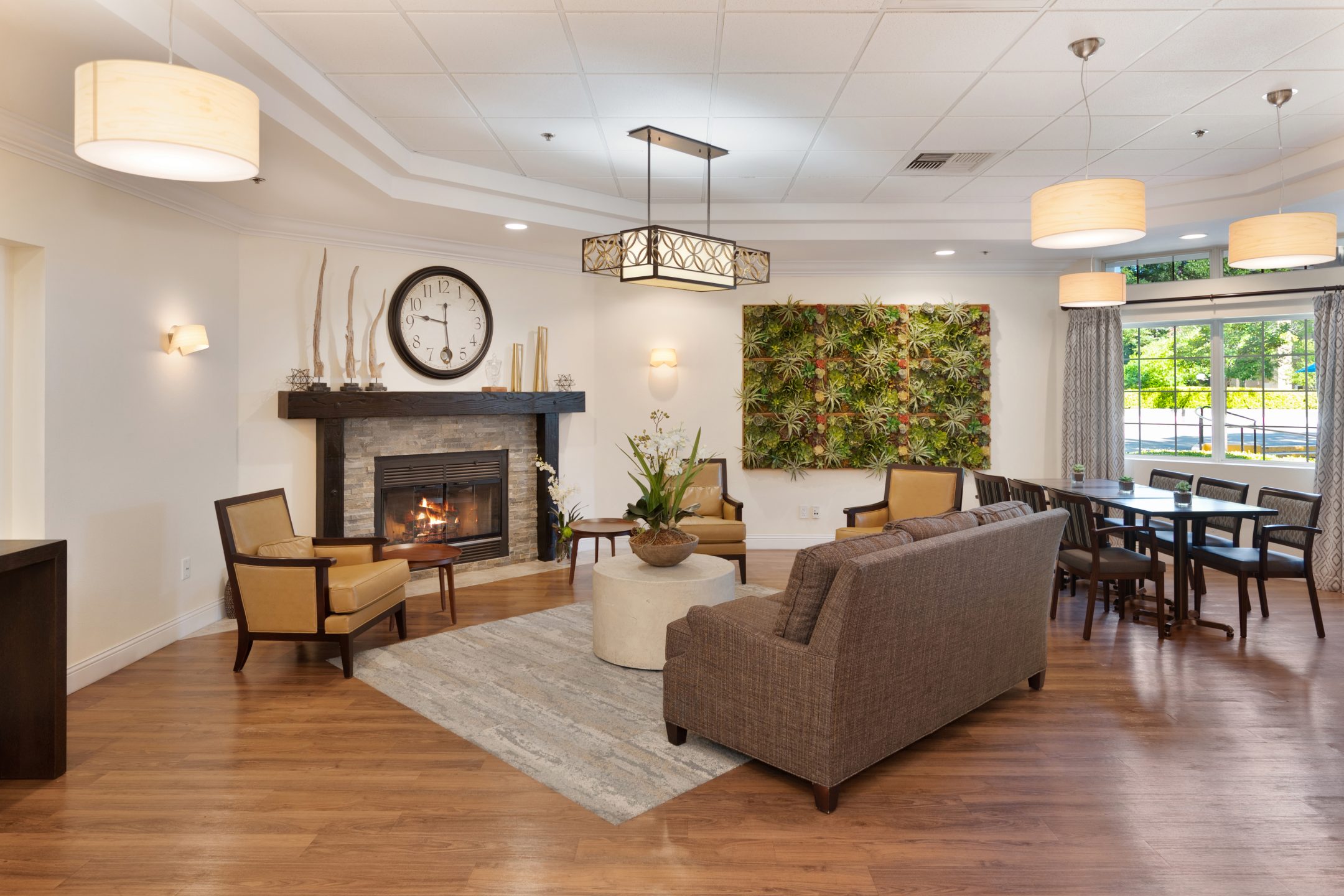 A modern, well-lit lounge perfect for Short-Term Stays, featuring a fireplace with decorative pieces on the mantel, armchairs, a sofa, dining table, and large windows overlooking lush greenery.