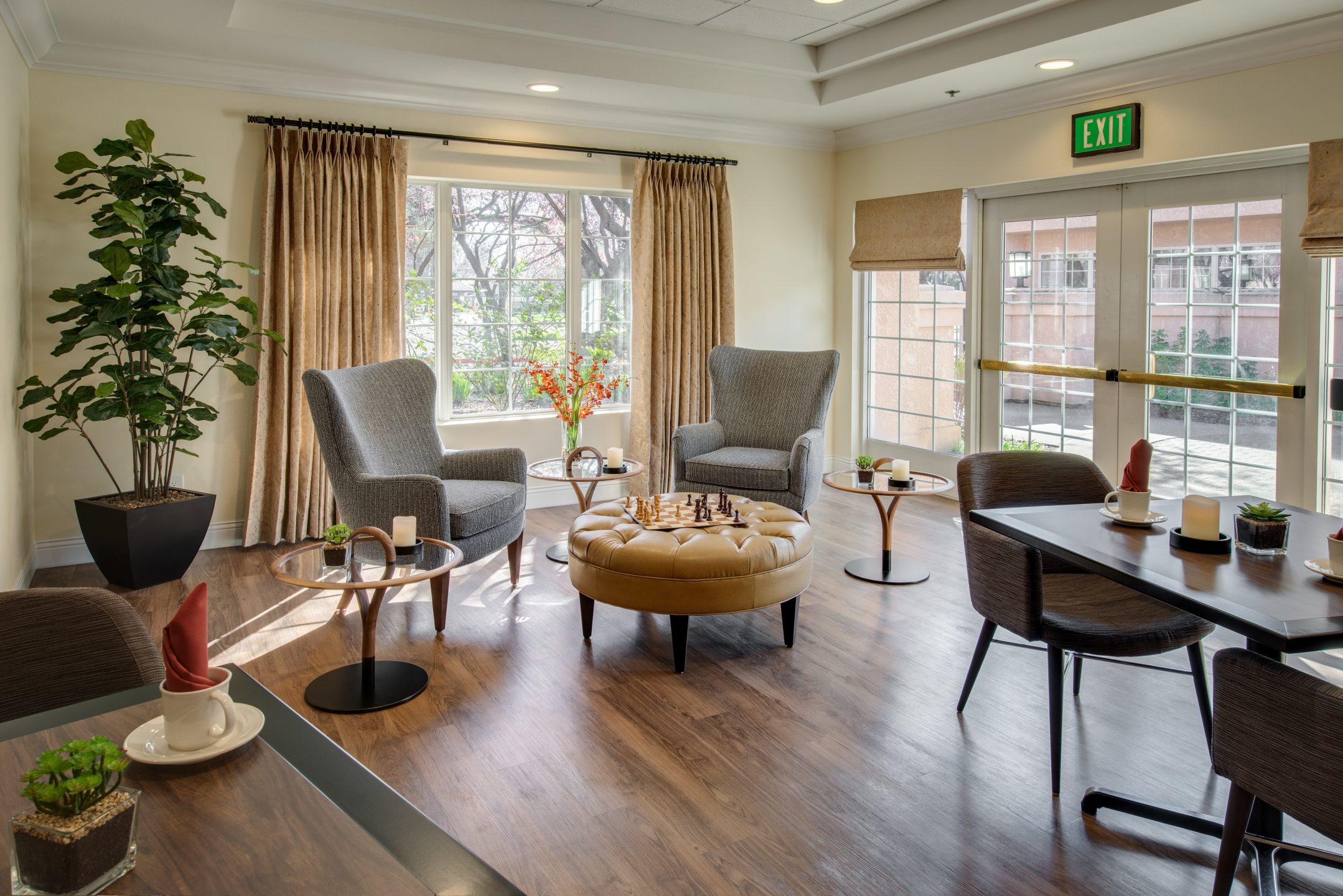 A cozy lounge with two gray armchairs near a window, a round ottoman with a chess set, and dining tables for two highlights inviting Living Options. Large windows and glass doors fill the space with natural light, creating a welcoming atmosphere.