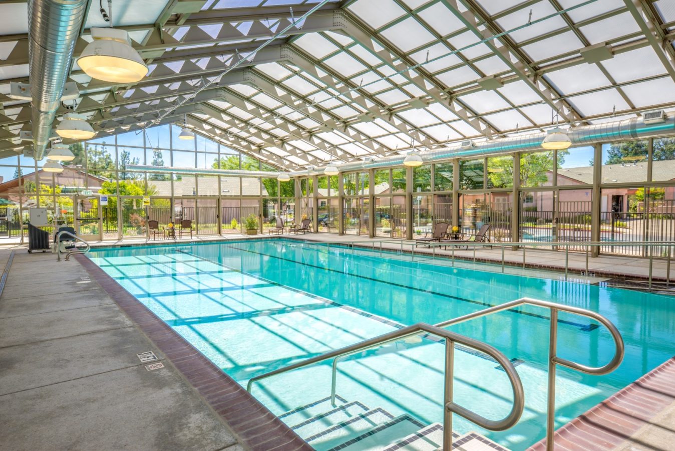 Bright indoor swimming pool with clear blue water, metal handrails, and large windows letting in natural light. Enjoy views of the outdoor seating and greenery—just one of many amenities offered in this inviting Life Plan Community.