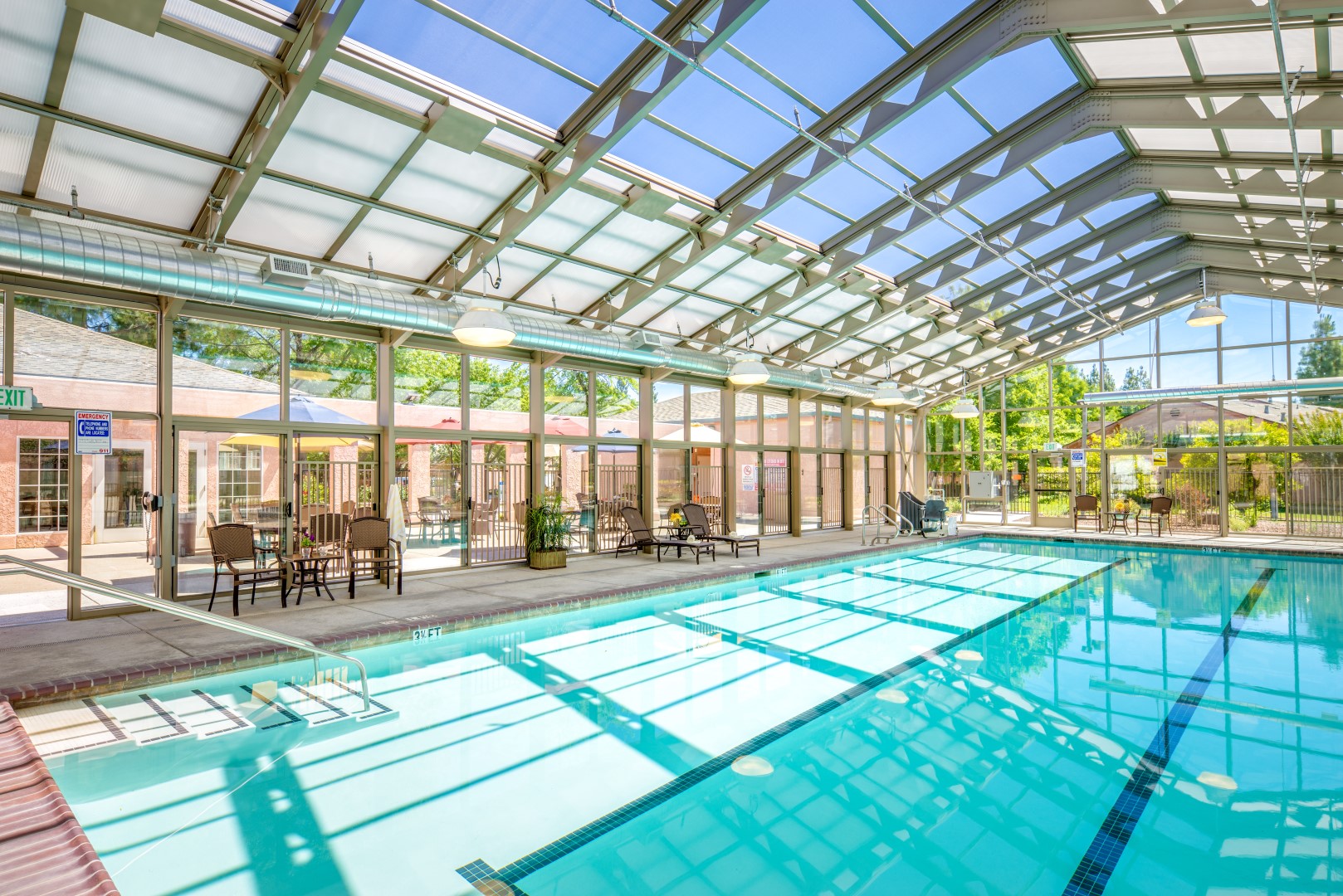 Indoor swimming pool with clear glass ceiling and walls, letting in sunlight. Poolside tables and chairs are set up along one side, and greenery is visible outside through the windows.