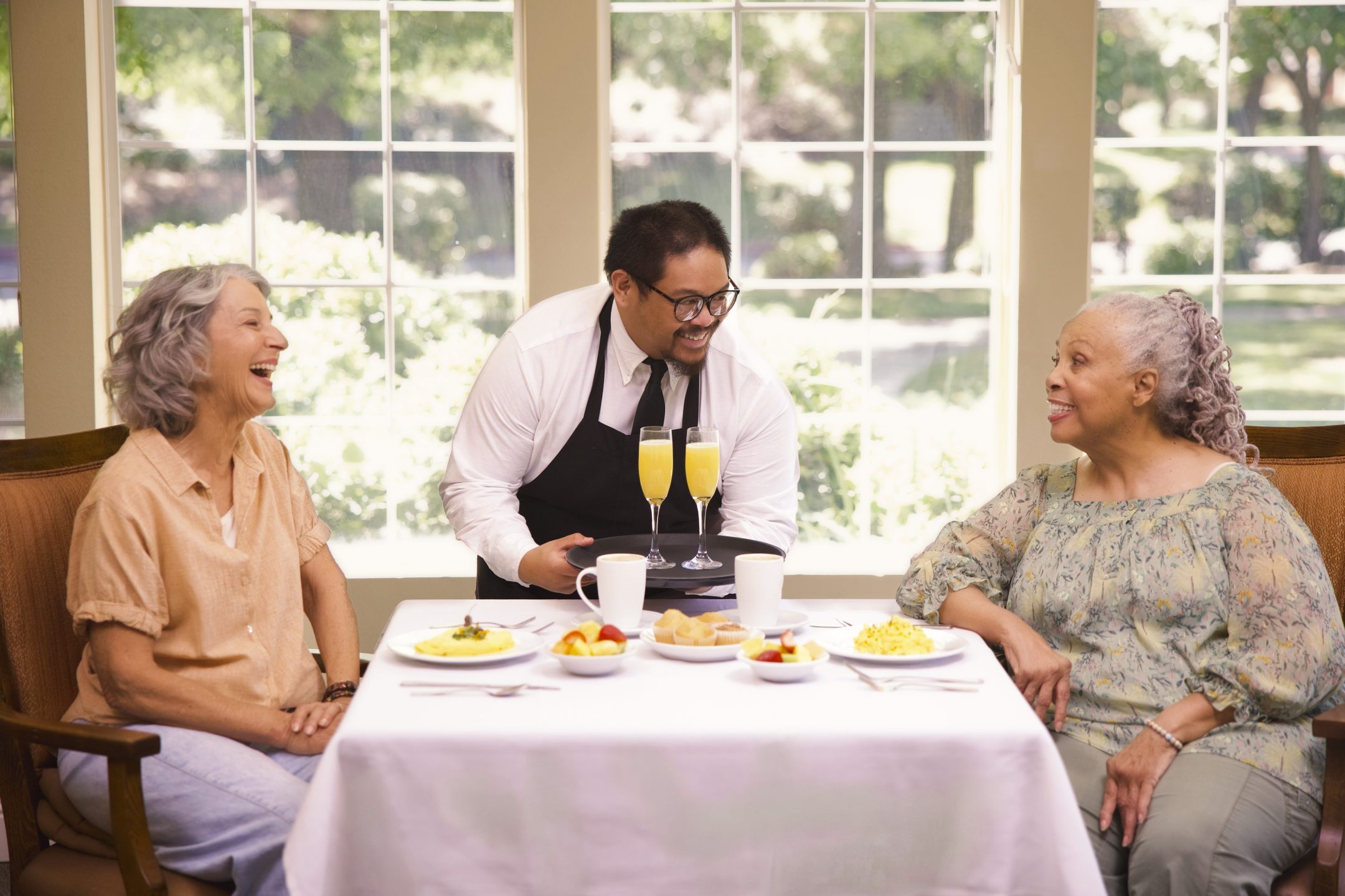 Two older women sit at a table enjoying a delightful culinary experience, smiling and talking with a waiter in a bright dining area with large windows and a garden view.