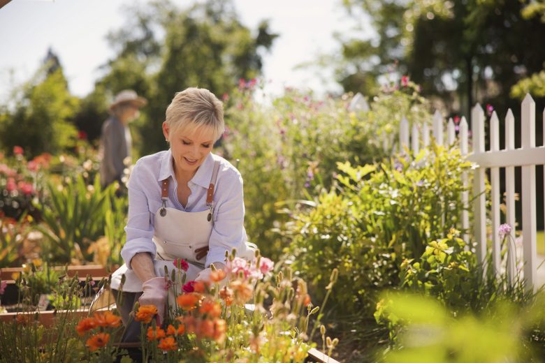 A woman with short blonde hair wearing a white shirt and apron smiles while tending to colorful flowers in a sunny garden, part of a skilled nursing rehabilitation program; a white picket fence and greenery are in the background.