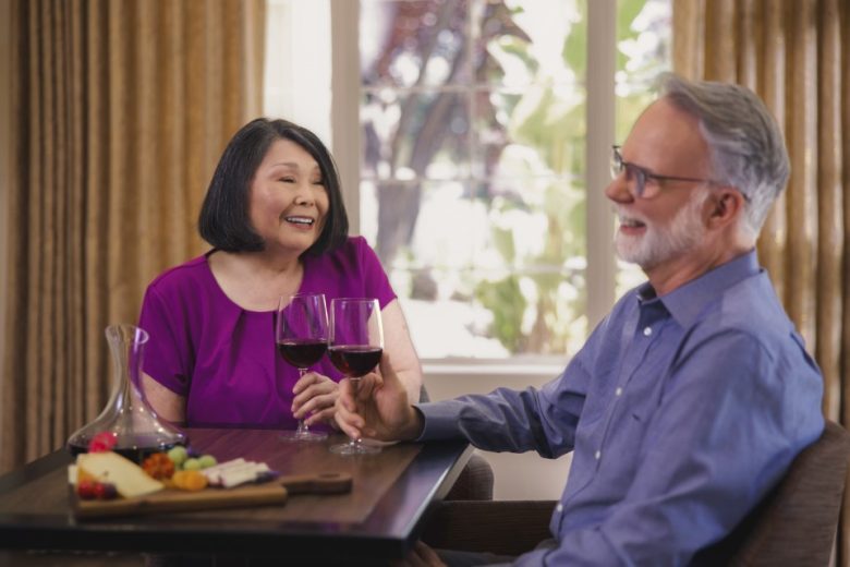 An older couple sits at a table indoors, smiling and holding glasses of red wine. There is a decanter and a wooden platter with cheese, fruit, and crackers in front of them. Sunlight filters through a window behind them.