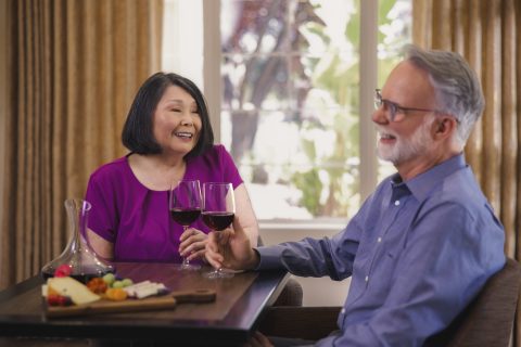 An older woman and man sit at a table, smiling and holding glasses of red wine. A wooden platter with cheese and fruit highlights their delightful culinary experience, while a window with curtains frames the cozy scene.