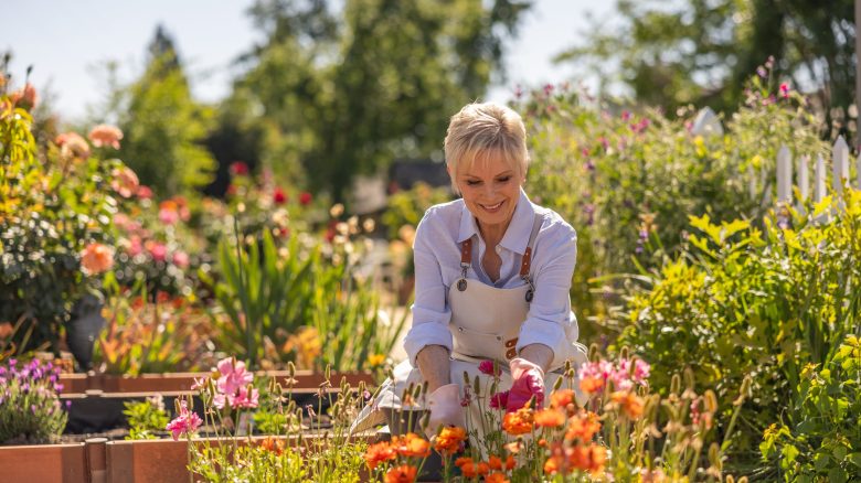 A woman wearing a white shirt and apron smiles while tending to colorful flowers in a sunny, vibrant garden at a Life Plan Community. Trees and more plants are visible in the background.