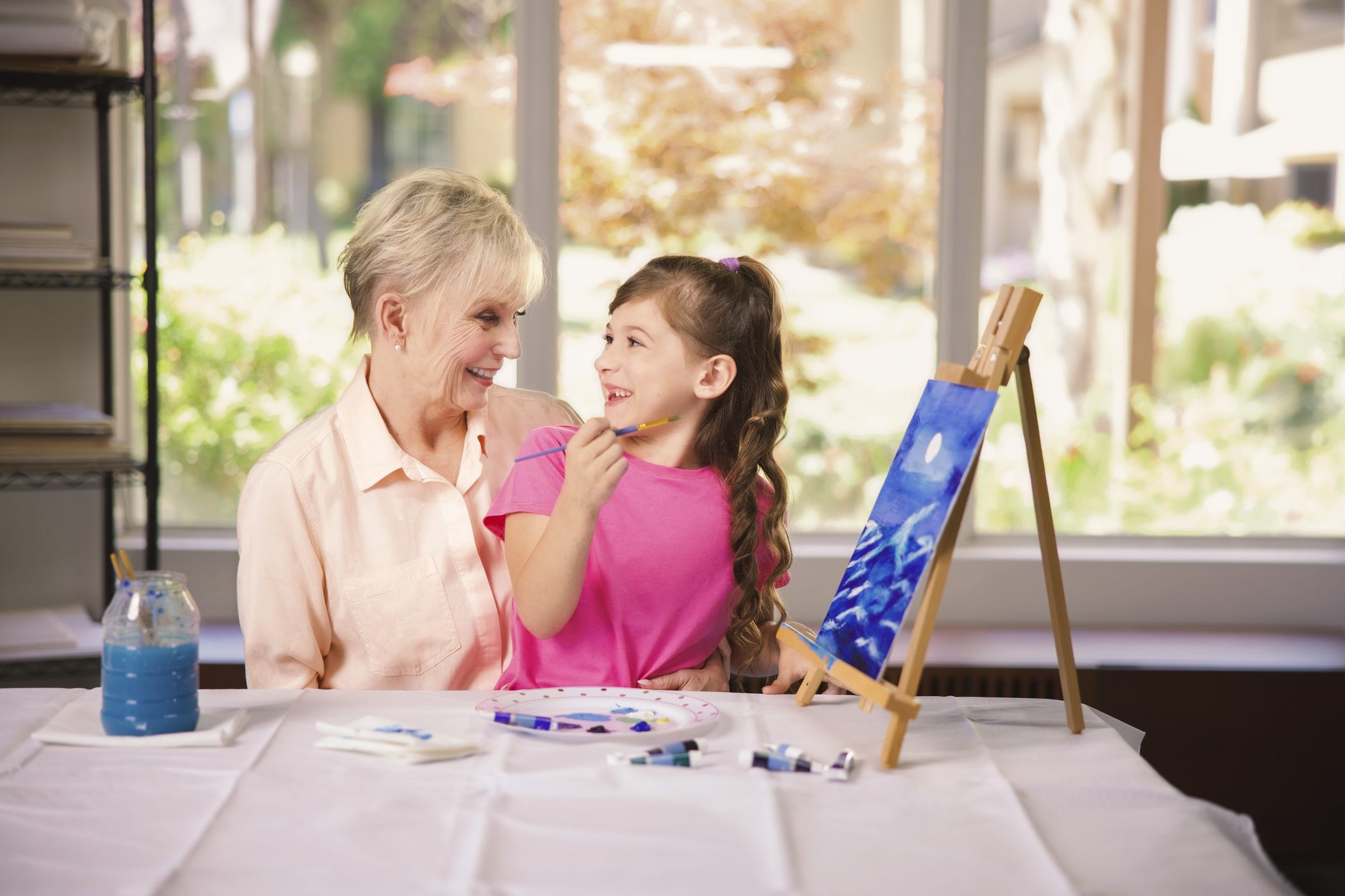 An older woman and a young girl smile at each other while painting together at a table, enjoying the warmth of resident culture. The girl holds a paintbrush, working on a blue canvas as sunlight streams through large windows behind them.