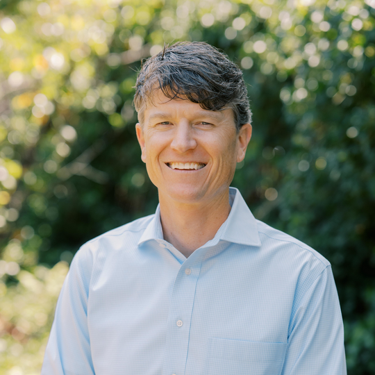 A man with short, wavy brown hair wearing a light blue collared shirt smiles while standing outdoors, with green foliage and sunlight in the blurred background.